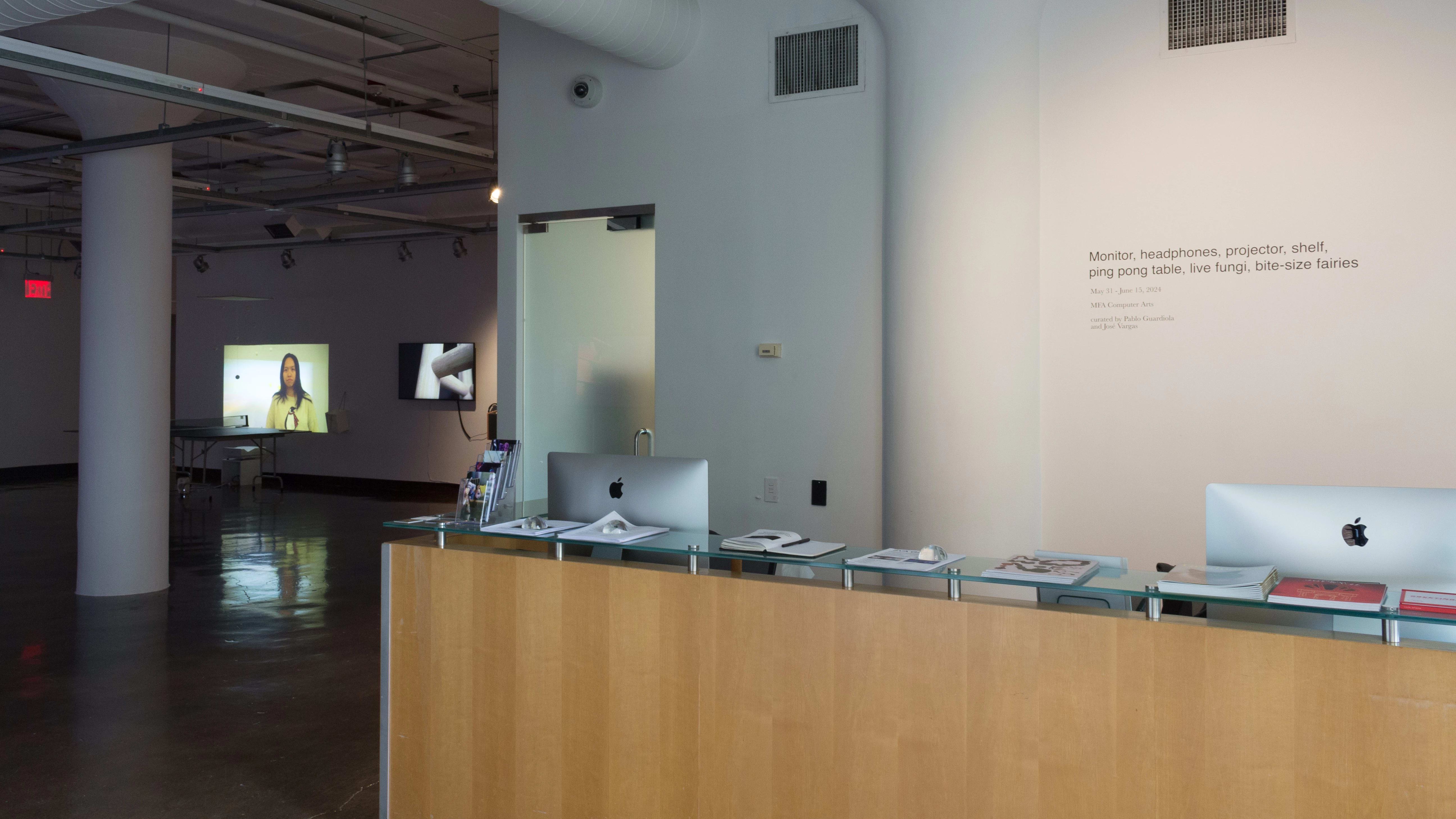 Installation image showing the title wall behind a computer desk, with a large gallery in the background to the left.