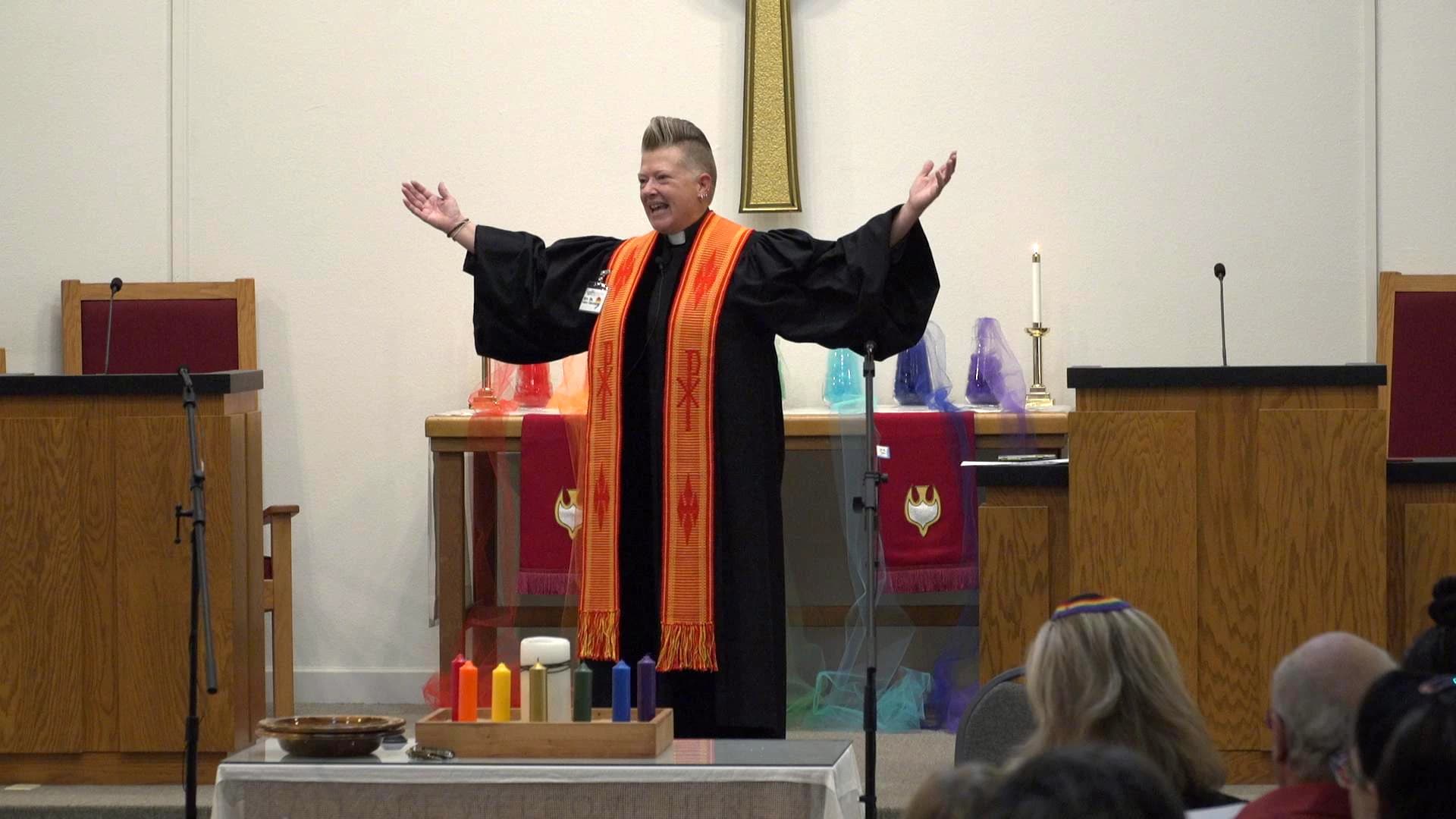 A priest dressed in black is inside Christian Church with his hands up and a smile on his face.
