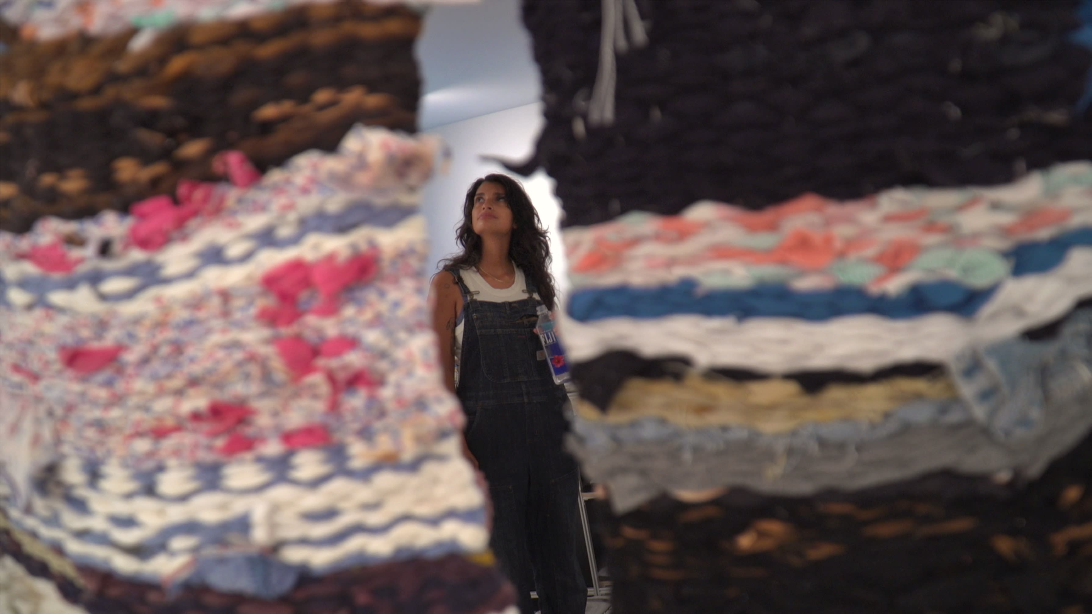 A female artist is standing in the center of two tapestries, looking at the tapestry she made.