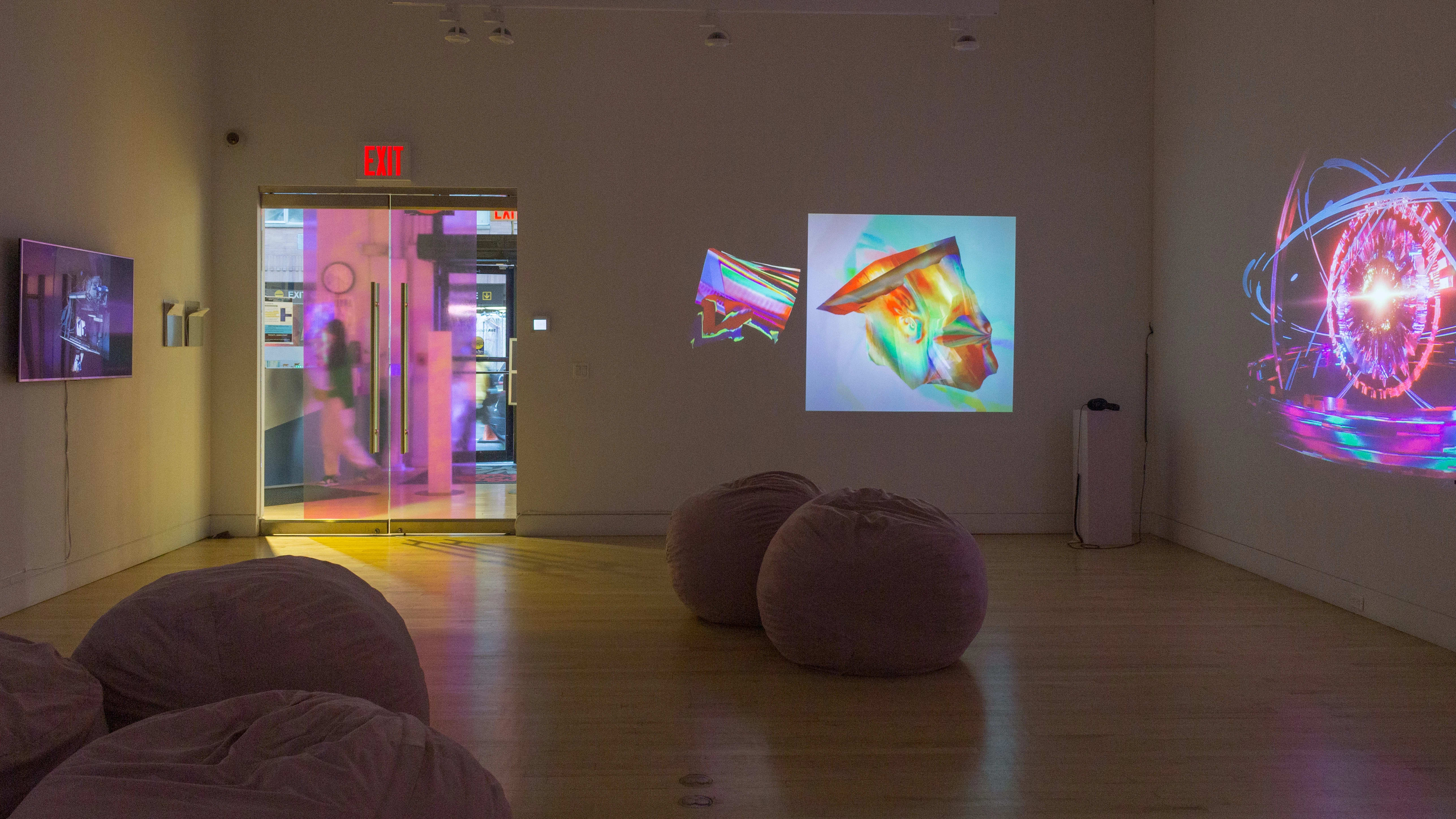 View from back of gallery space looking toward the entry door, with a monitor playing an artwork on the far left, a diptych of abstract projected artwork in the center, and on the far right wall is an animation of a brightly-colored universe. 