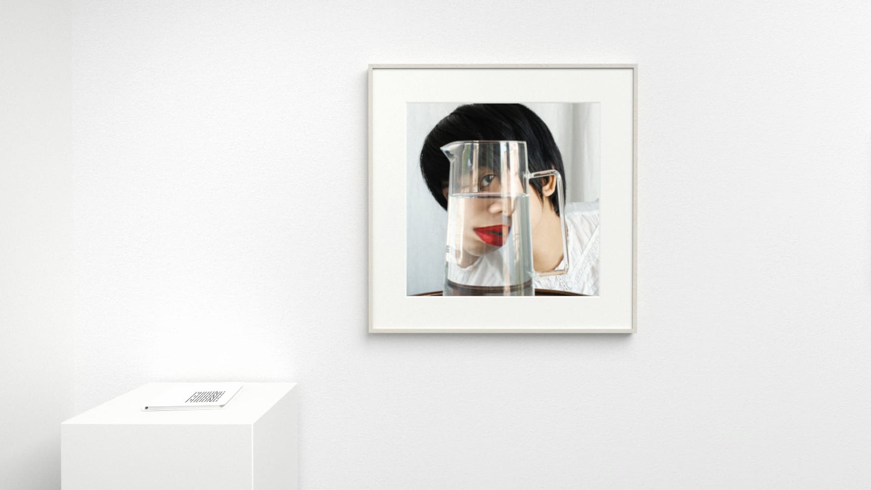 Color photograph of a person looking through a partially filled pitcher of water, in white frame, and alongside it is small white book on a pedestal