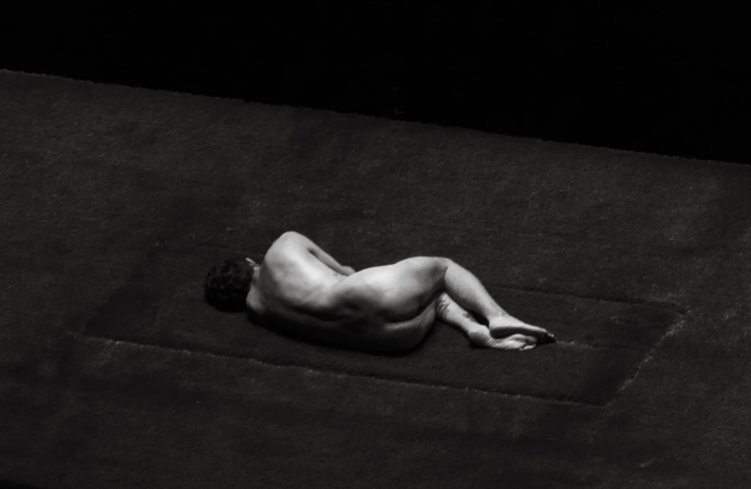 black and white image of naked man from behind in curled position, laying on grass. 