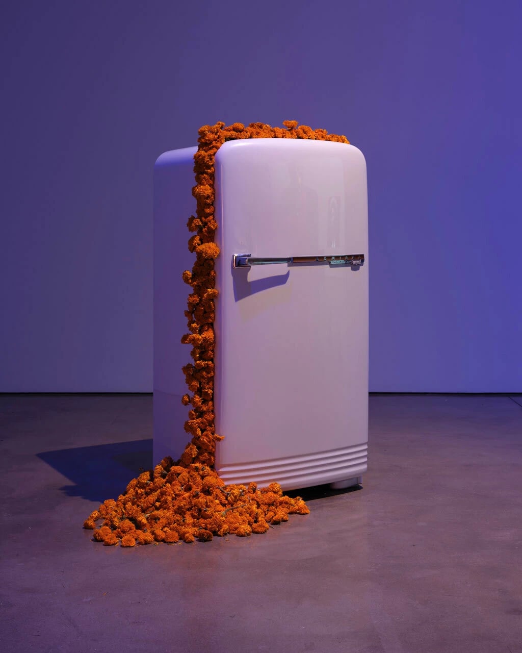 A picture of a refrigerator, with its doors mostly closed but with a large amount of orange flowers bursting through the crack and scattering across the floor.