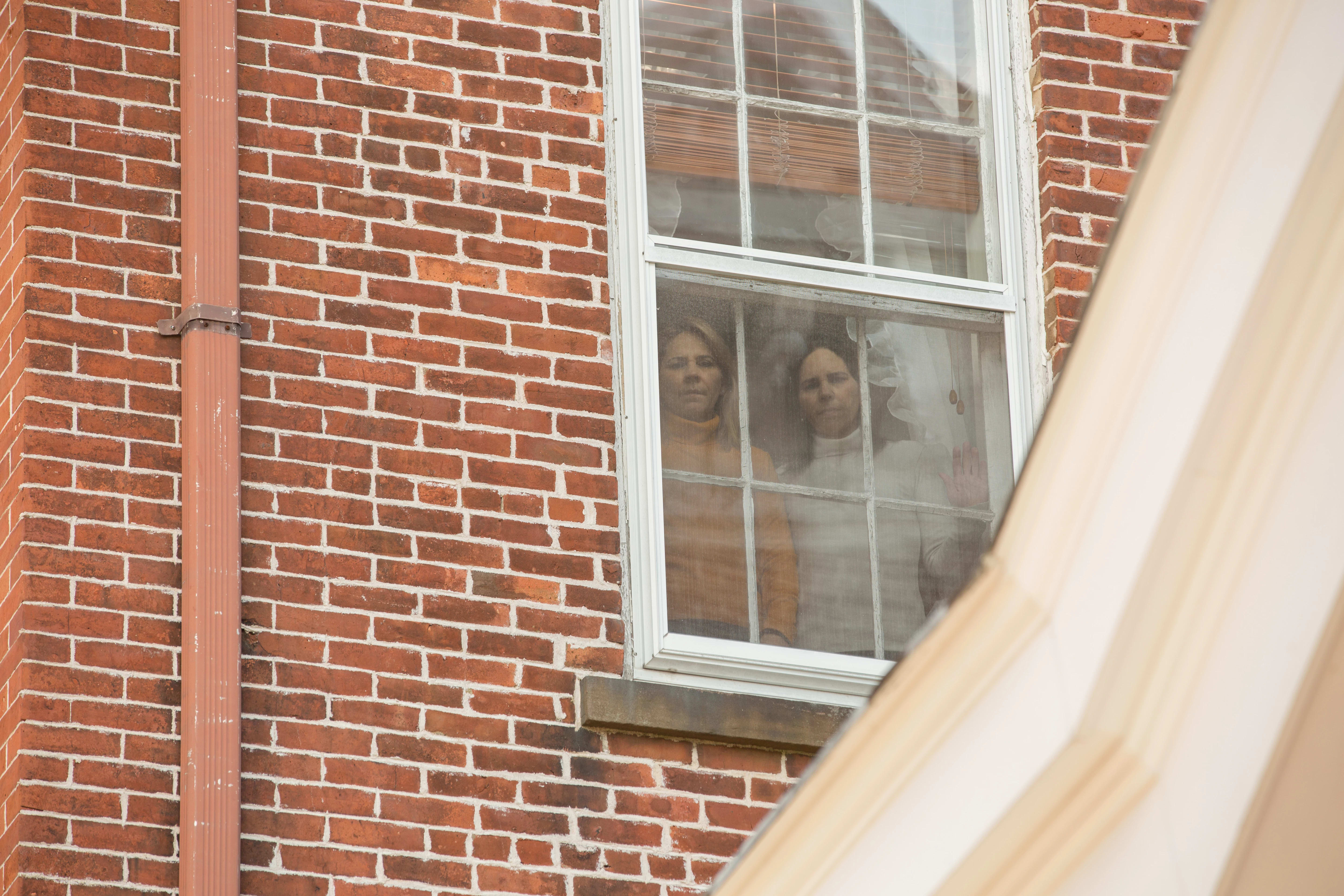 Photograph was taken outside of a brick wall house where you can see two women standing in front of the window one wearing yellow mustard turtle neck shirt and the other women is wearing a white turtle neck shirt and they are both standing next to each other looking straight to the camera.