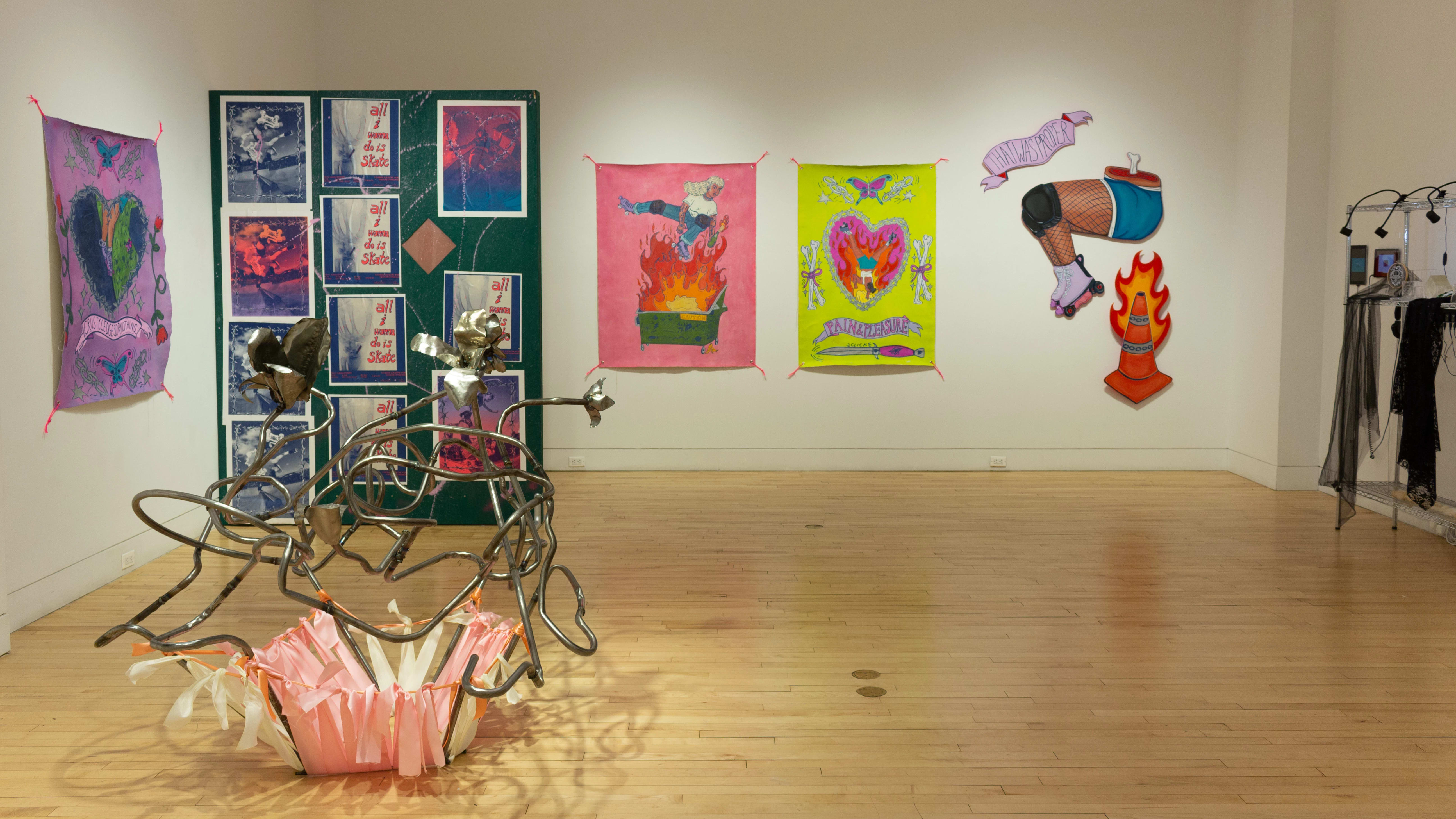 Installation image featuring a wire sculpture on the floor in the foreground, multiple fabric prints ion the wall, a large painted wooden wall with prints in the background, paintings depicting human legs with skates, and a metal rack.