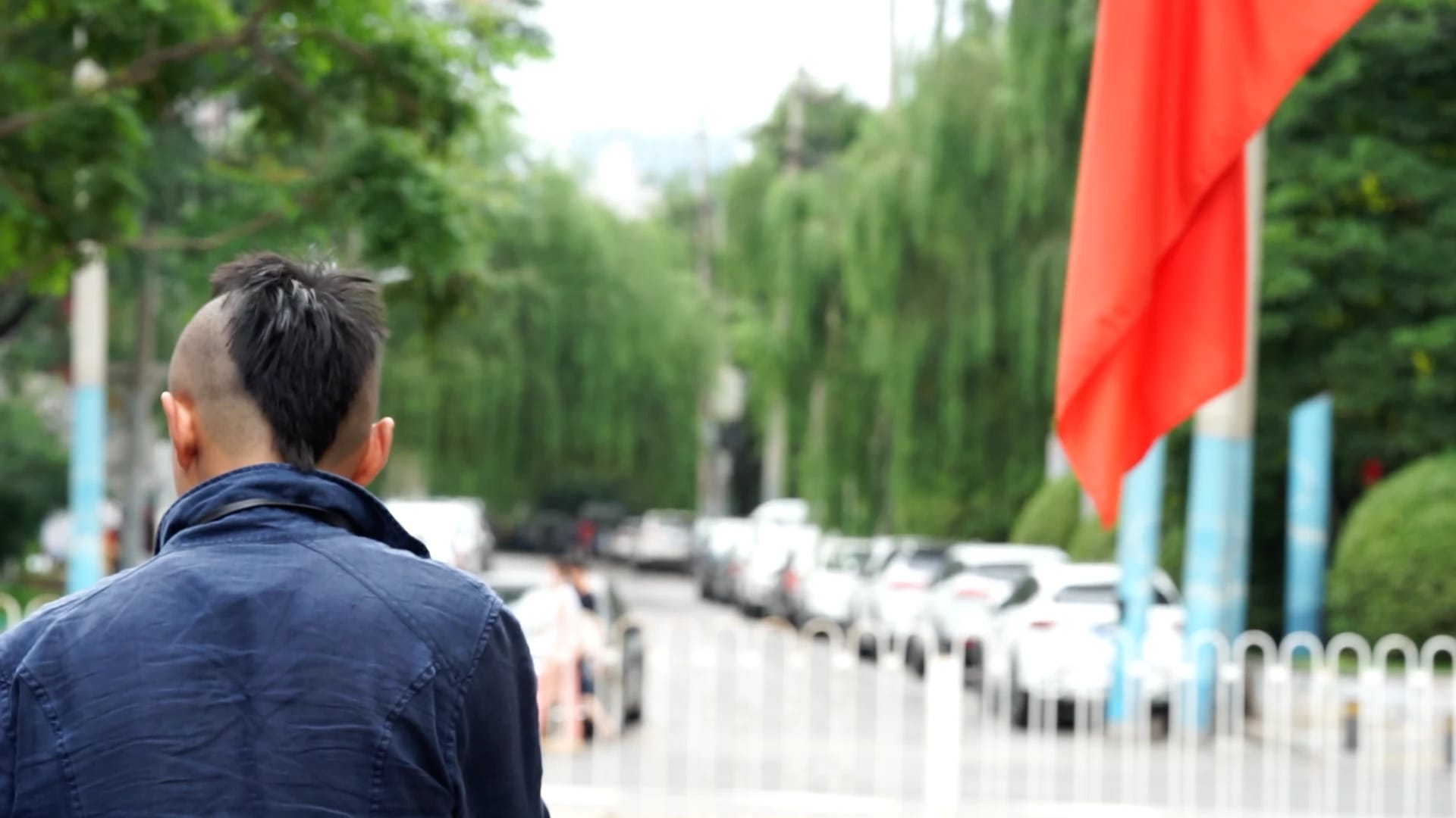 Half back of a young man walking down a Beijing street with Chinese red flags and a row of willow trees on the side of the road.