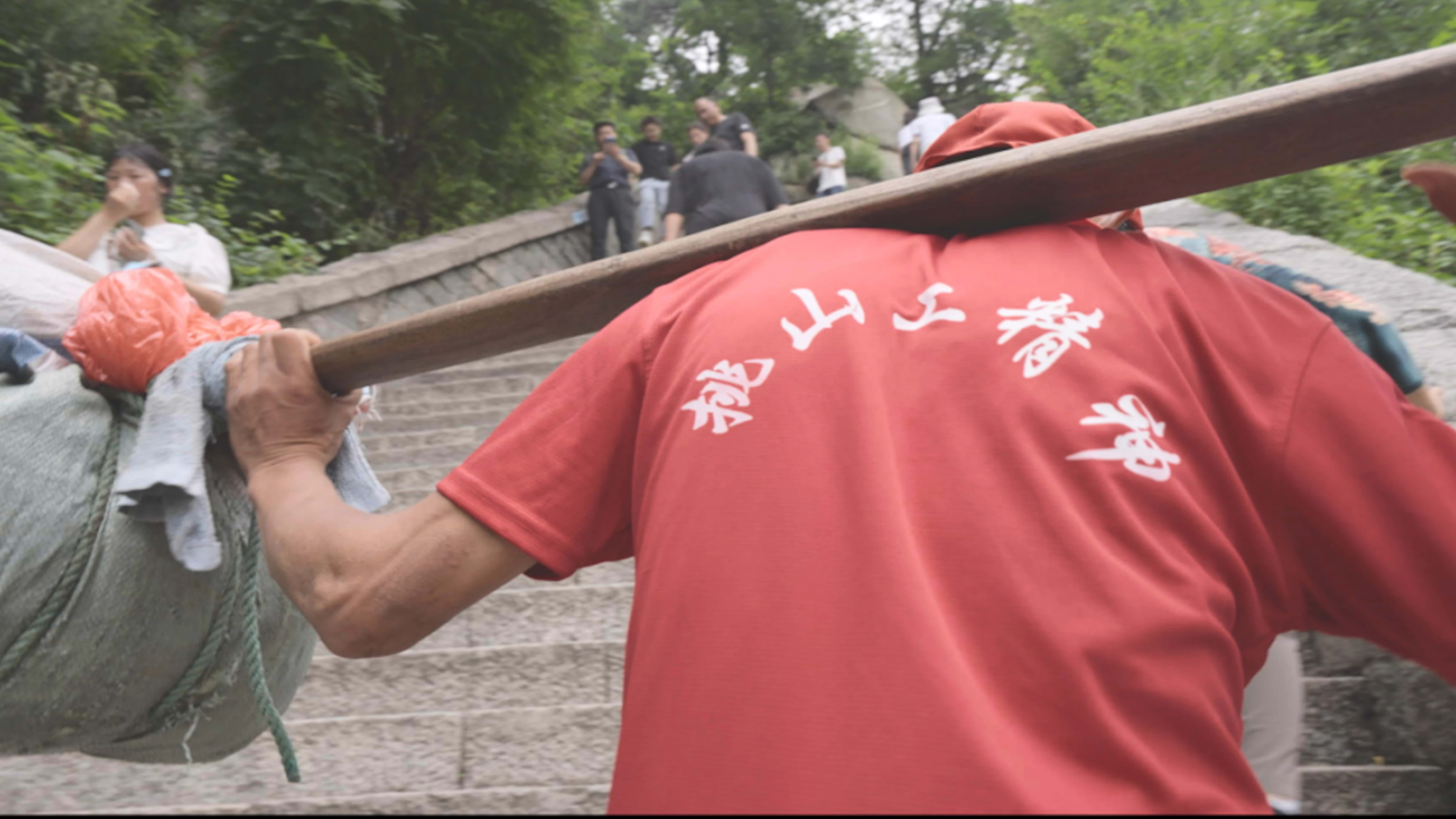 The back of a man dressed in red as he walks forward on the steps of China's Mount Tai, carrying a heavy load on a stretcher.