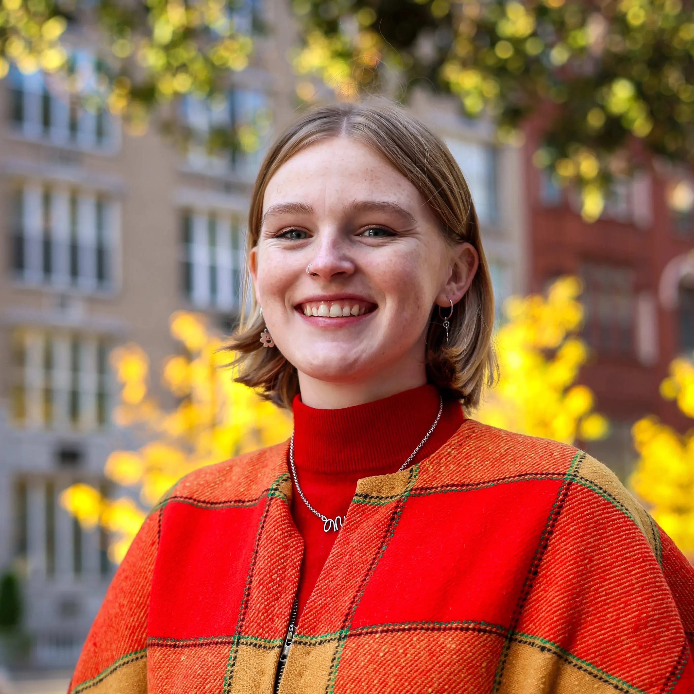 A woman with dark blonde hair smiling at the camera. She's wearing a red turtle neck, a red plaid outer layer and a thin metal necklace