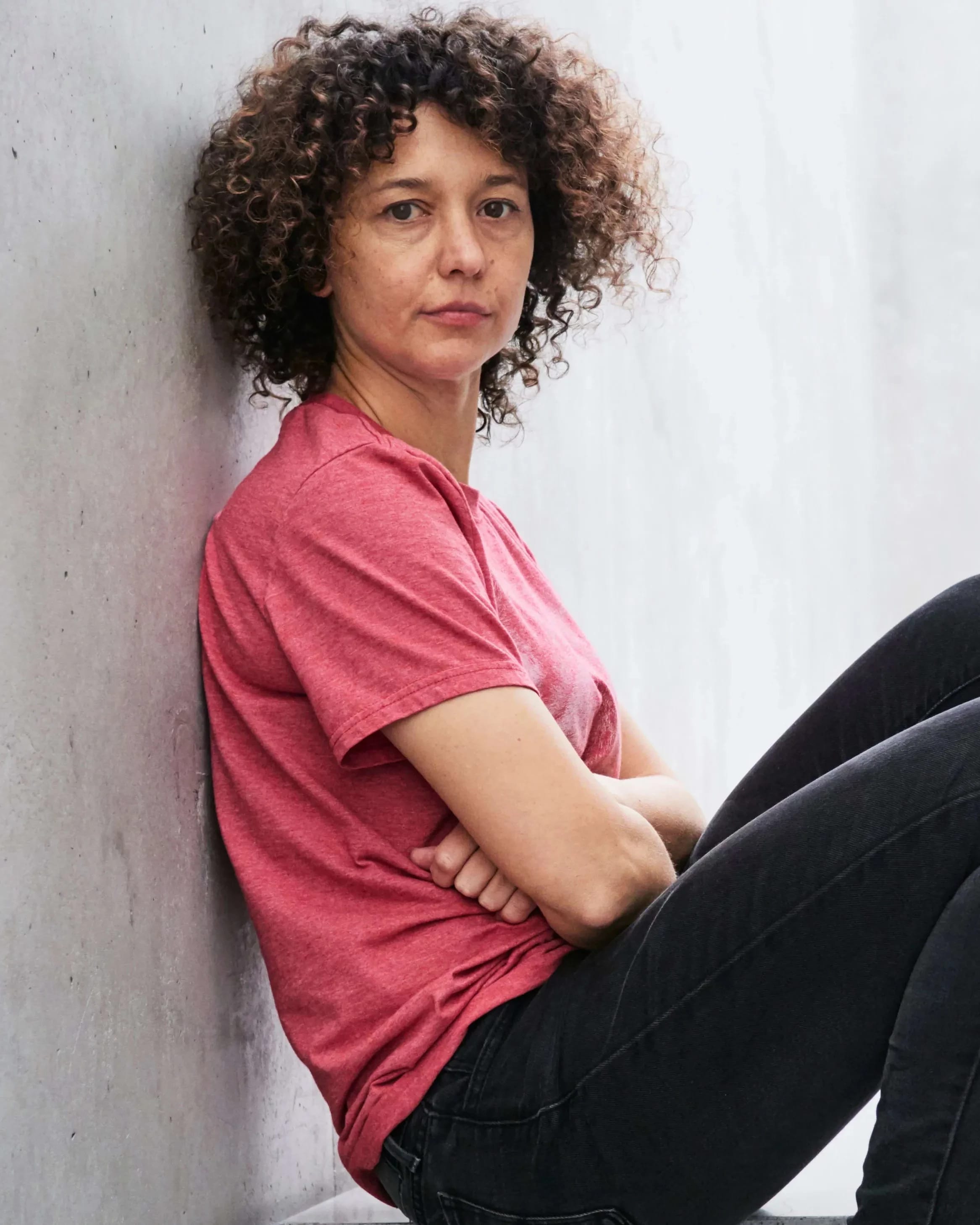A photograph of Mika Rottenberg. Mika is sitting on a cement step with her back against the wall. She is wearing a red t-shirt with black jeans and boots. Her head is turned to the camera with a relaxed expression.