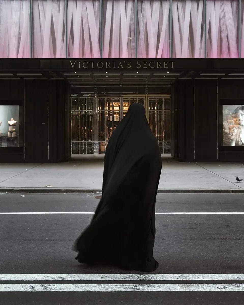 A color photograph of a figure enveloped in a flowing black garment walks across a city street, directly in front of a large Victoria's Secret store. The storefront features large, illuminated advertisements of models in lingerie on either side of its main entrance.