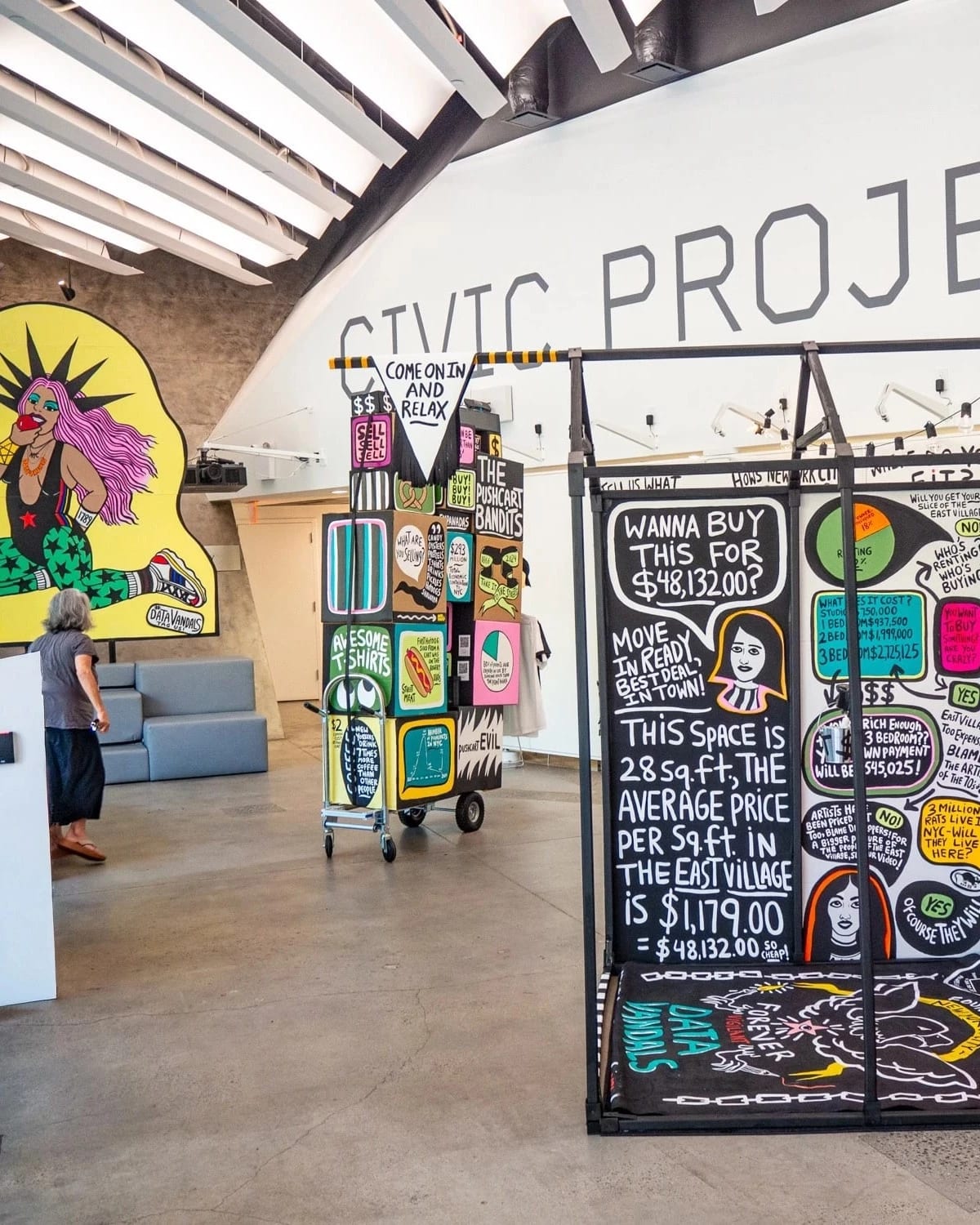 Interactive data visualization installation in a modern gallery space featuring colorful, street art-inspired panels with housing cost data. The central black panel displays NYC housing statistics including '$48,132.00' and text about East Village rental prices per square foot. Surrounding panels feature vibrant graphics with pink, yellow, and green elements. A person in casual clothing examines the display while standing on polished concrete floors under industrial ceiling architecture.