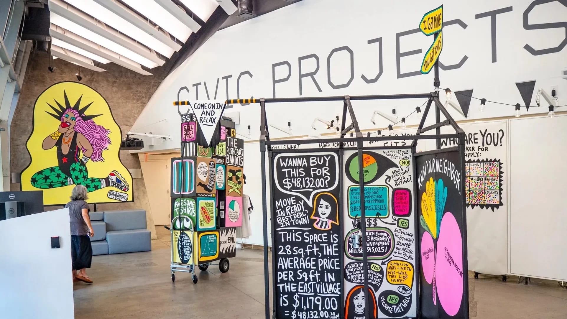 Interactive data visualization installation in a modern gallery space featuring colorful, street art-inspired panels with housing cost data. The central black panel displays NYC housing statistics including '$48,132.00' and text about East Village rental prices per square foot. Surrounding panels feature vibrant graphics with pink, yellow, and green elements. A person in casual clothing examines the display while standing on polished concrete floors under industrial ceiling architecture.