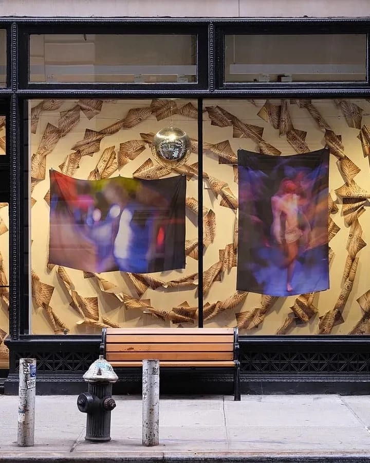 Large windows displaying two abstract fabric artworks and a disco ball inside; a wooden bench and fire hydrant are on the sidewalk in front.