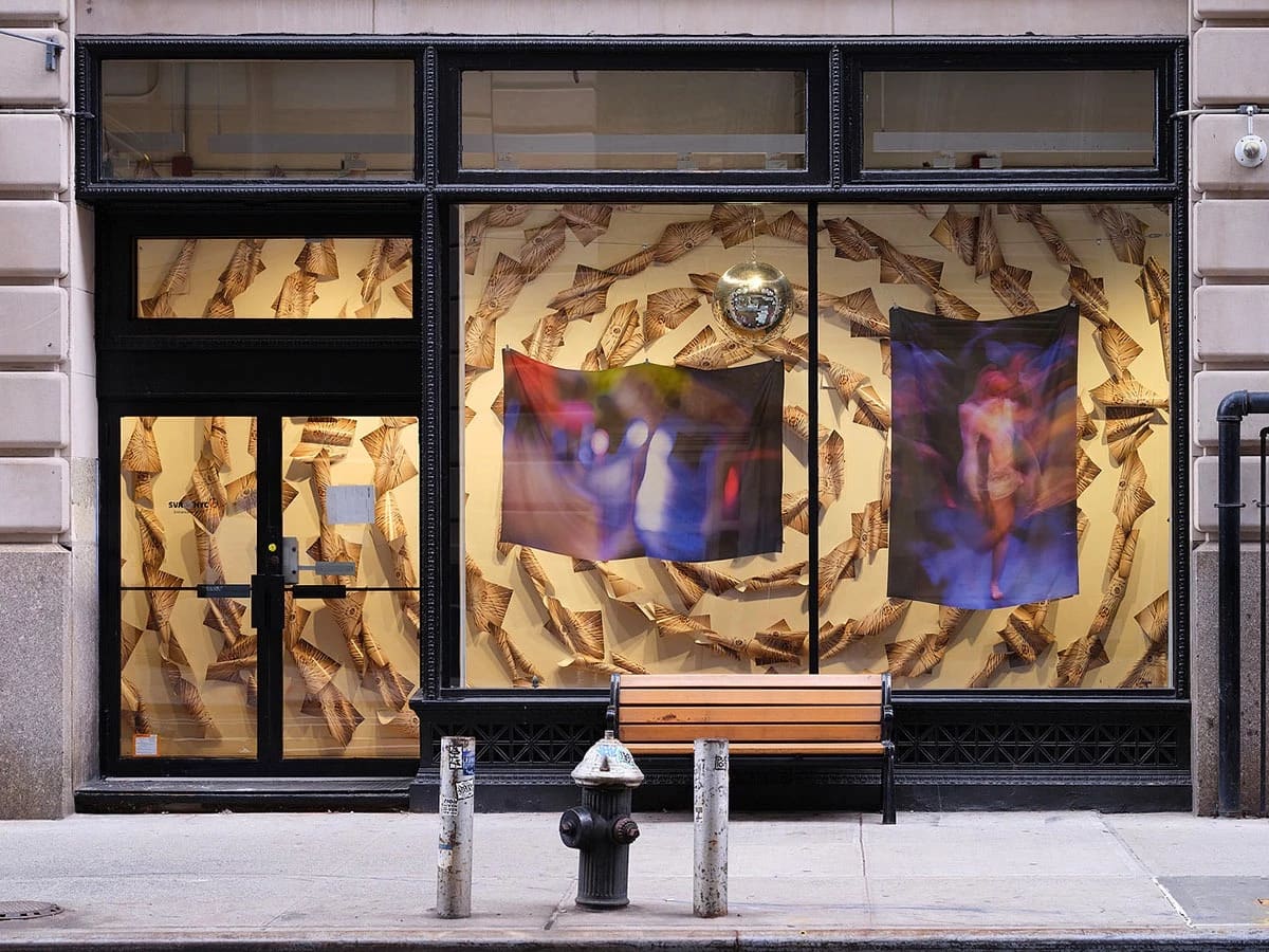 Large windows displaying two abstract fabric artworks and a disco ball inside; a wooden bench and fire hydrant are on the sidewalk in front.