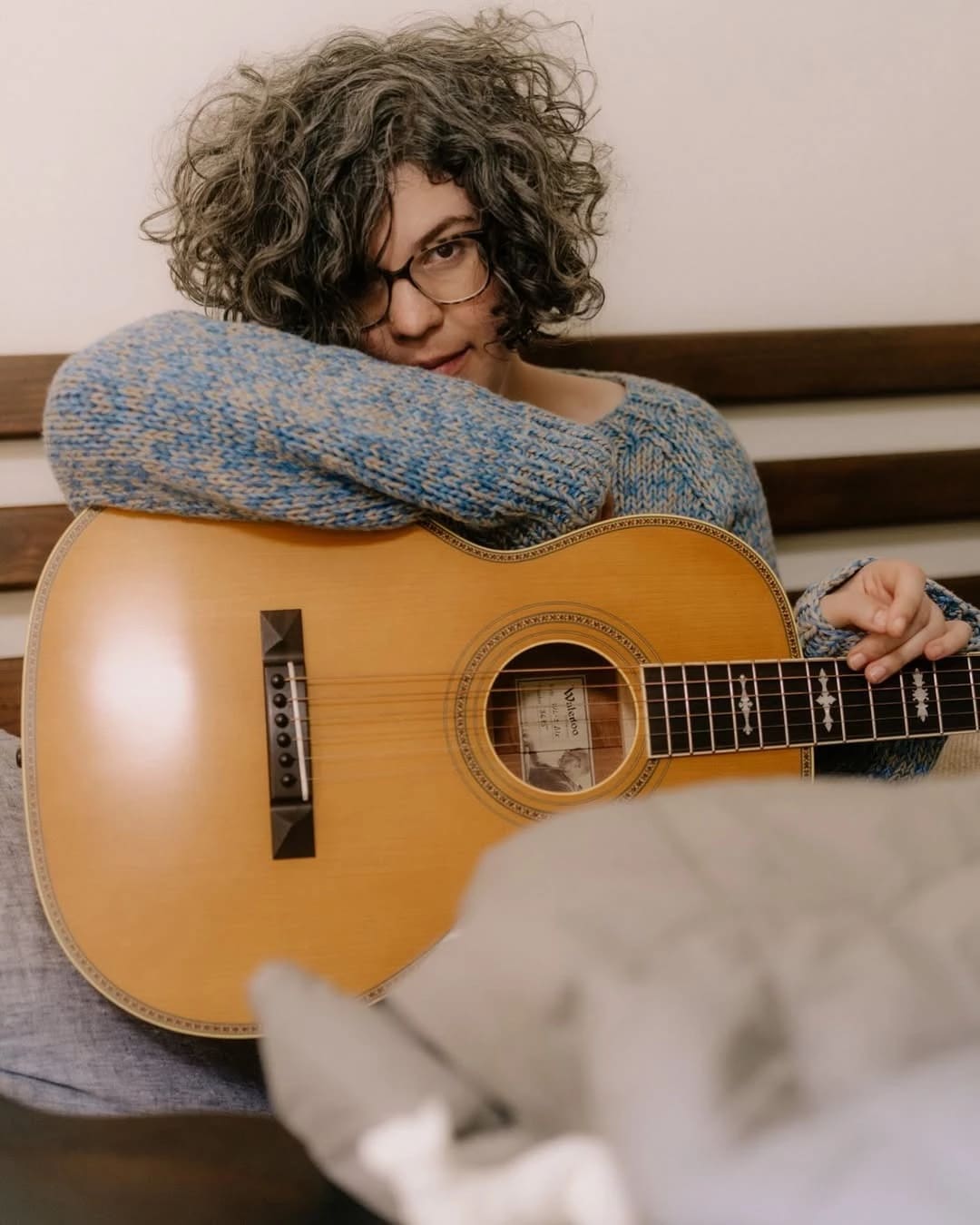 A person with curly hair and glasses leans on an acoustic guitar, wearing a blue sweater. The mood is relaxed and introspective.