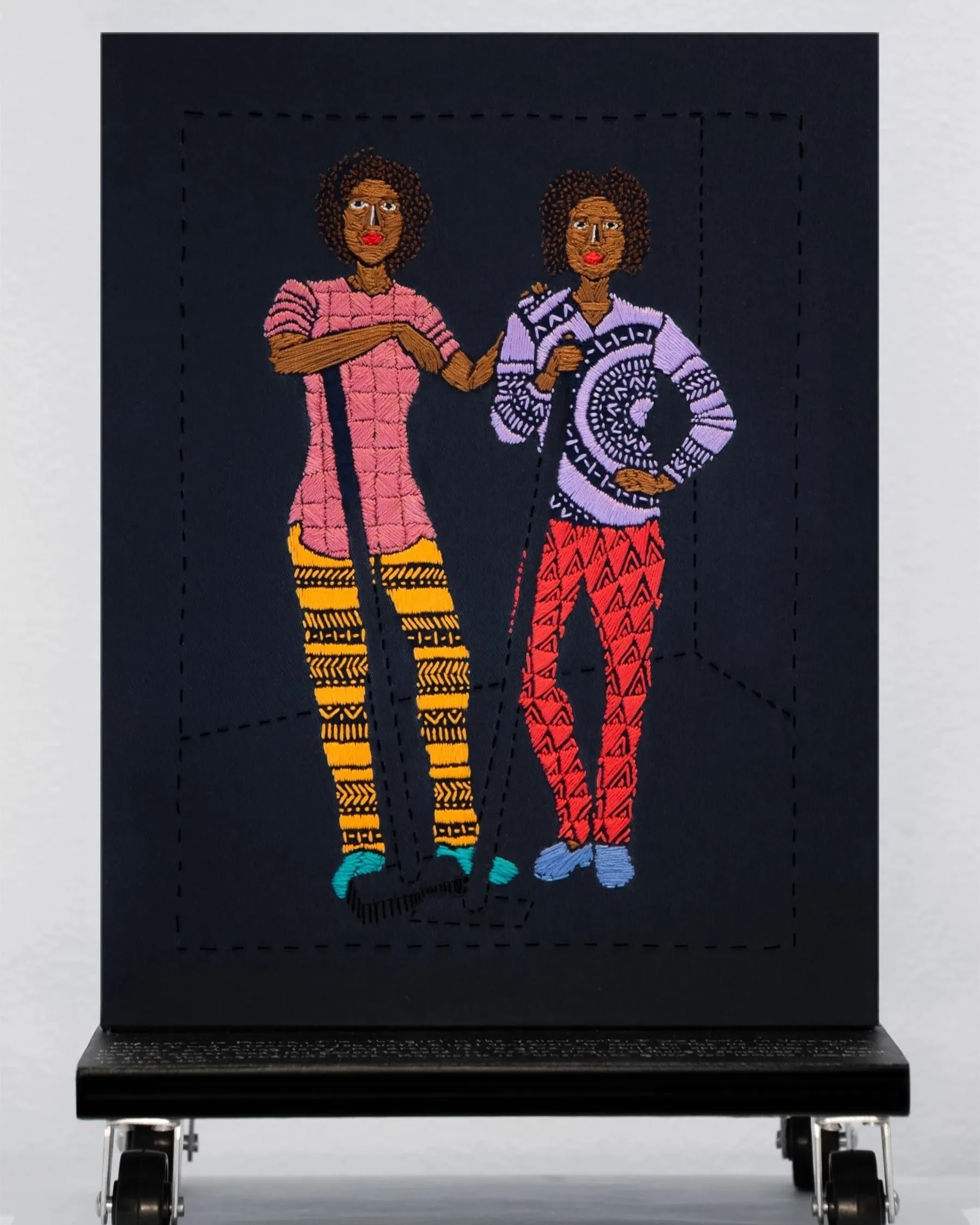 A black embroidered canvas with two figures of women standing and holding cleaning supplies.