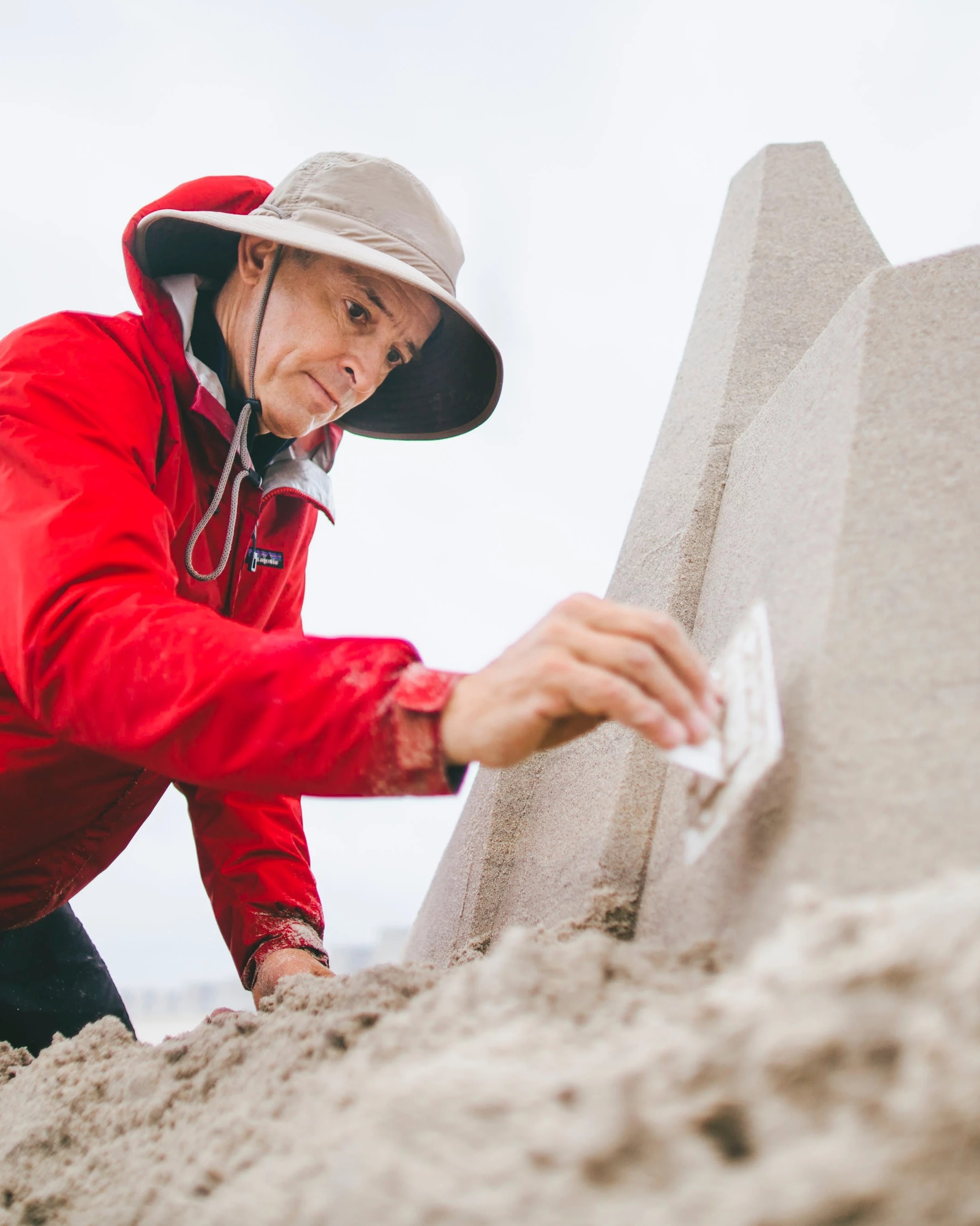 A man in a raincoat, hat, and shorts, kneels in the sand on a beach and works on a pyramidical sandcastle. He is tamping the sand down on one wall of the structure using a rectangular piece of plastic.