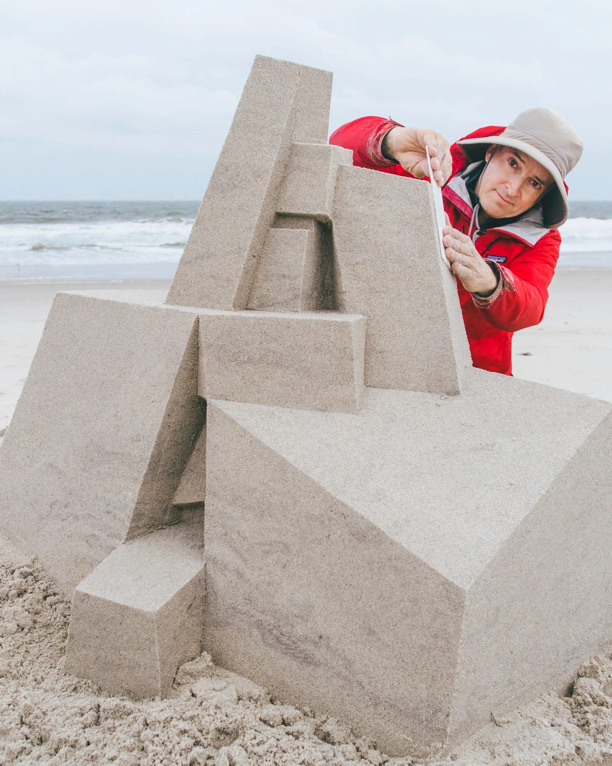 A man in a hat and raincoat on an empty beach puts the finishing touches on a sandcastle sculpture.