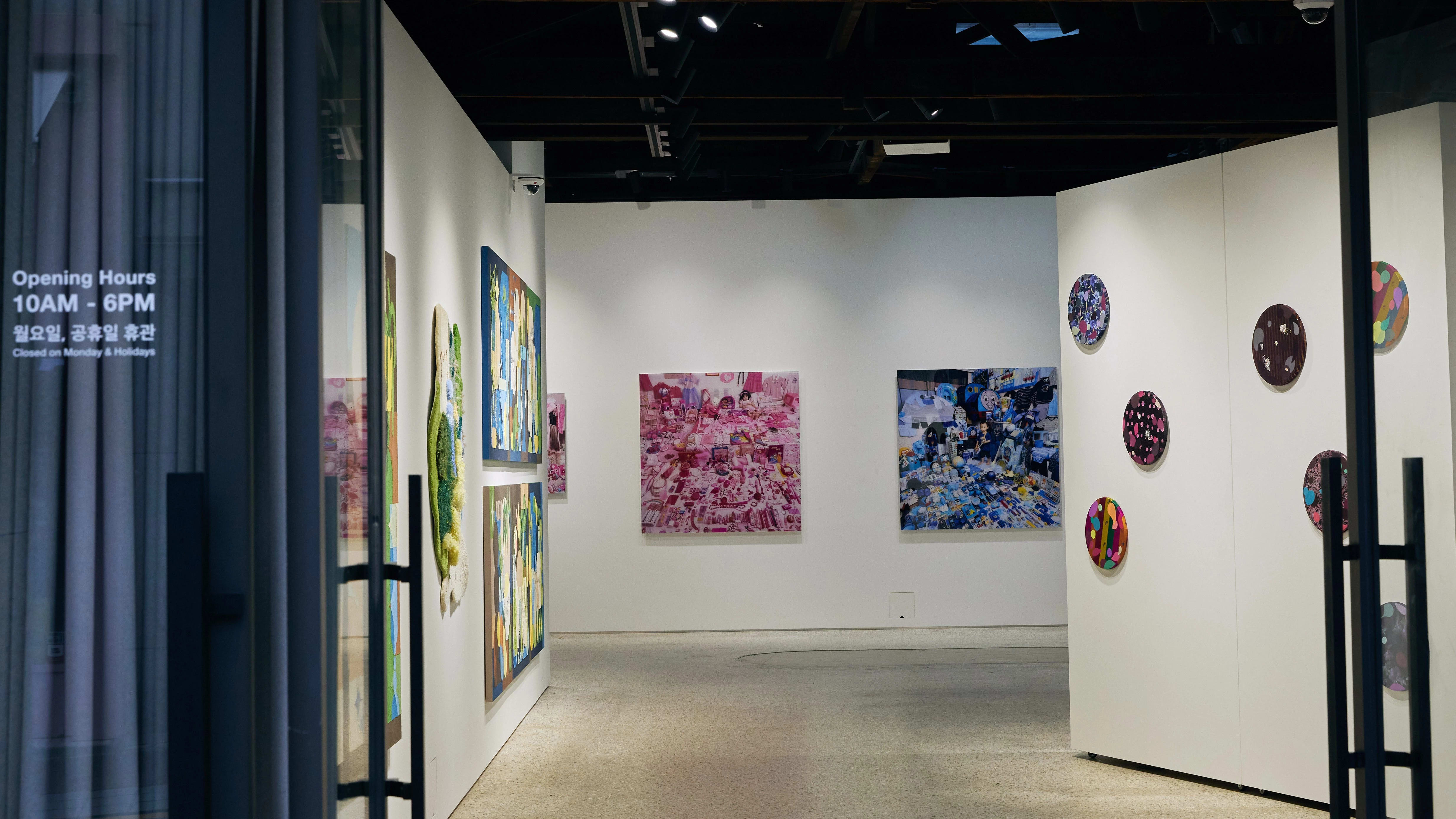 Doors propped open at a gallery's facade, revealing an in-progress exhibition with many art pieces hanging from the three visible walls.