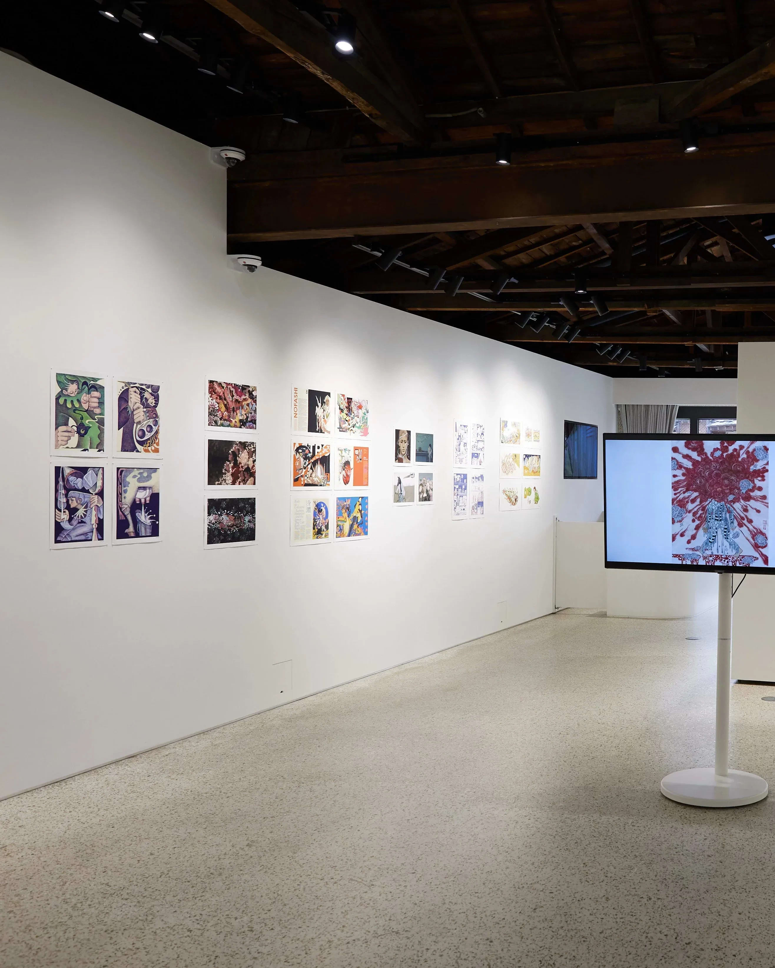 Gallery installation displaying colorful comic and illustration-style artworks arranged linearly along both walls. A large monitor on a white pedestal at the center shows a figure with red abstract patterns. The space features an open ceiling with exposed dark wooden beams, track lighting, and natural light filtering through glass doors at the far end.