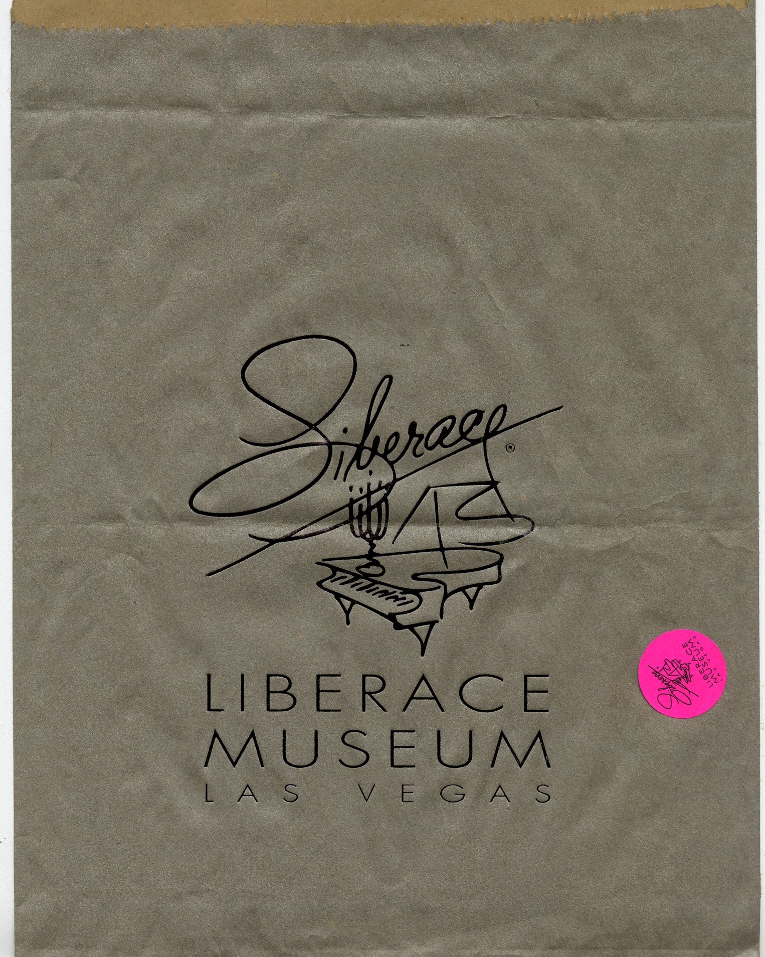 A brown paper back with the logo of the Liberace Museum Las Vegas. A fine line illustration of a piano with "Liberace" written in cursive handwriting on top. There is a pink sticker at the corner of the paper bag with the same logo but much smaller. 