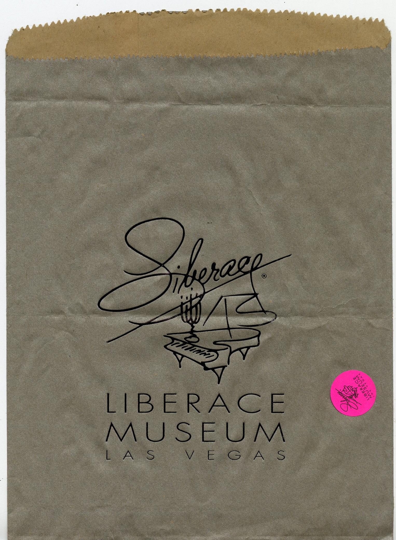 A brown paper back with the logo of the Liberace Museum Las Vegas. A fine line illustration of a piano with "Liberace" written in cursive handwriting on top. There is a pink sticker at the corner of the paper bag with the same logo but much smaller. 