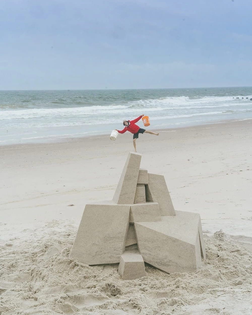 A man in a raincoat, hat, and shorts carrying a large bucket in each hand poses for a photo on an empty beach. In the foreground is a large sandcastle, and the man is posed so that he appears to be standing on the top of the sculpture.