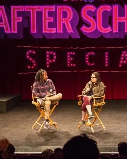 Two people sit on a stage in directors chairs in front of a curtain with the words "After School Special" in pink behind them. There's a crowd facing the stage in the foreground.