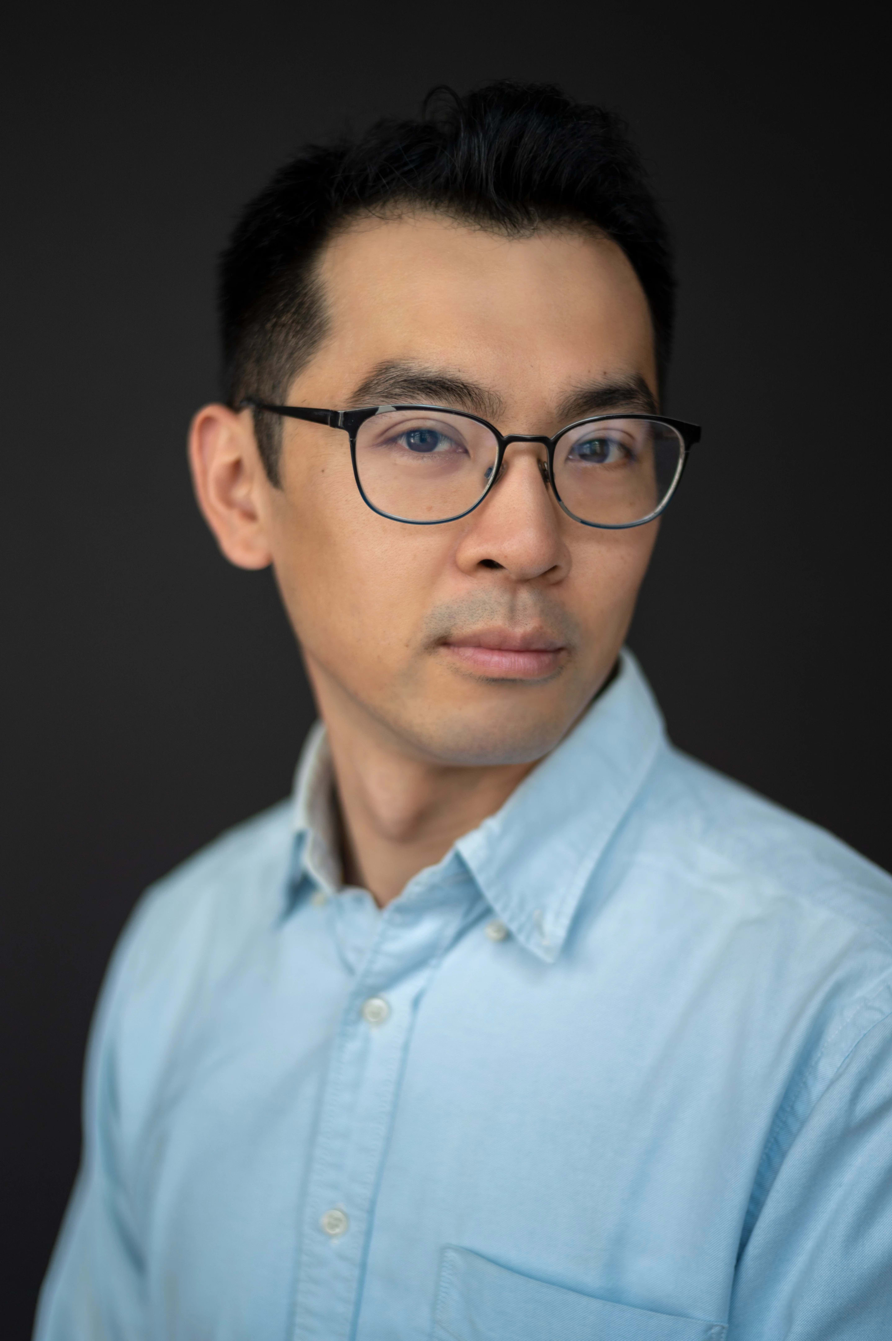 A photo of a man looking at the camera with his head turned to his left. He is wearing a blue shirt and glasses.