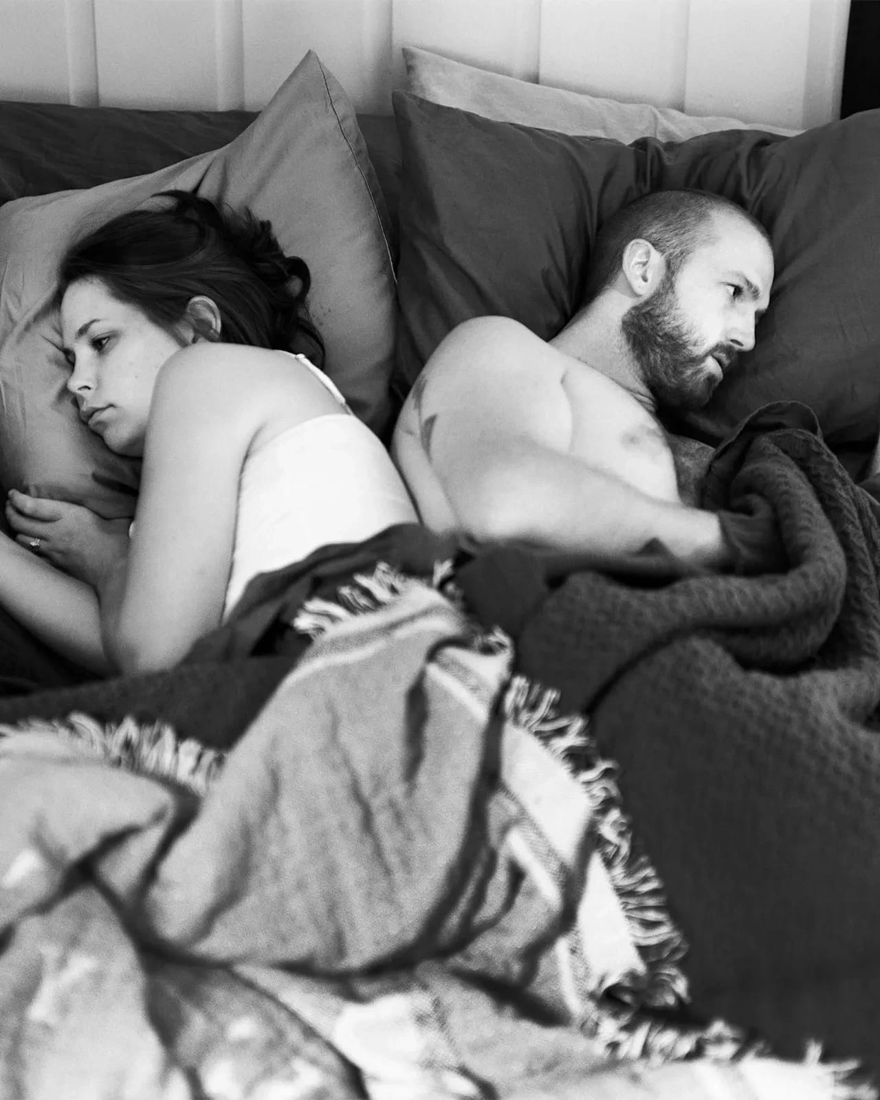 A black and white photograph of a man and a woman lying side by side in bed, facing away from each other. Each holds a hand in the air as if scrolling or holding a phone, though no devices are present. Their expressions are distant and disconnected. The photo is black and white and emotionally charged.