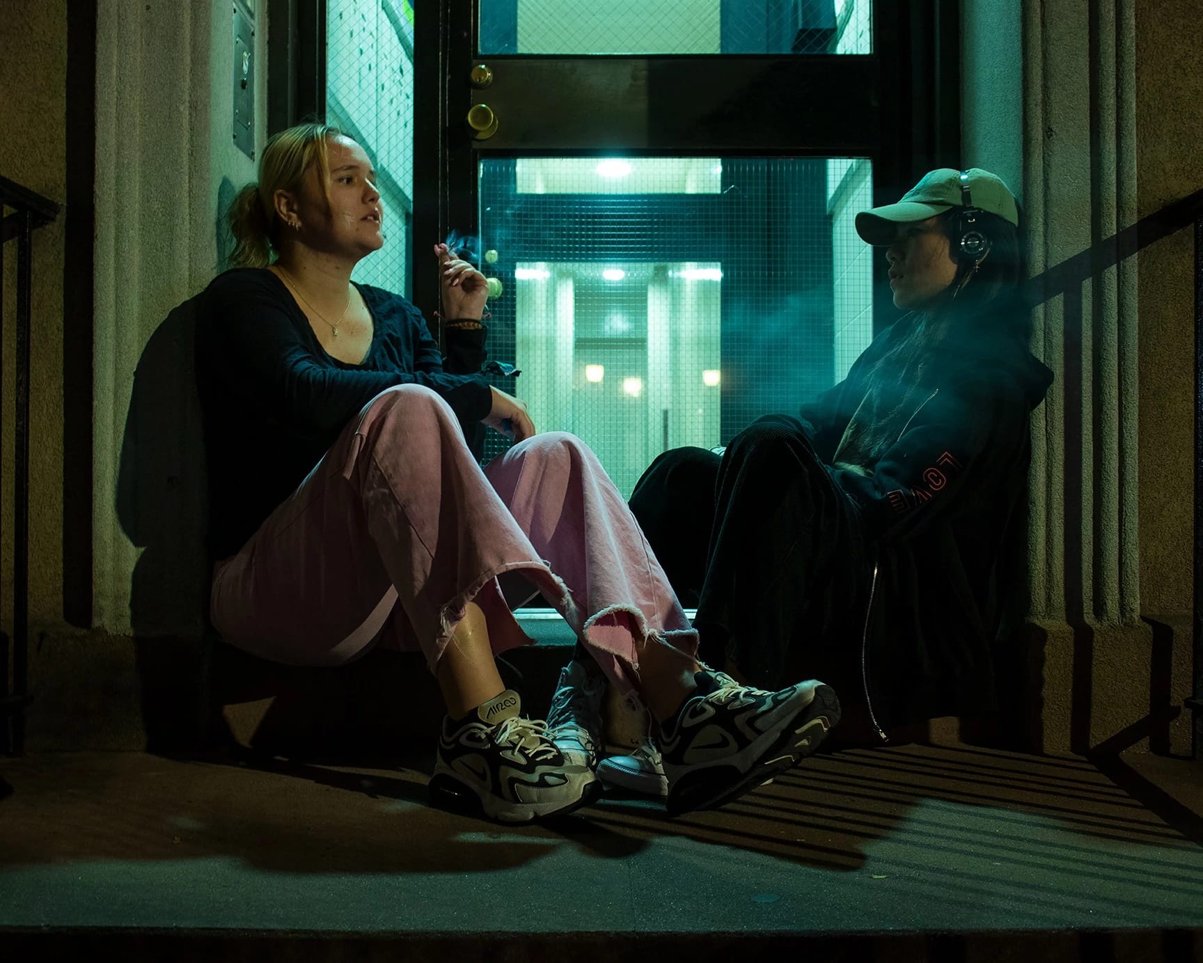 Image of two young women sitting outside on an apartment building stoop, one is smoking