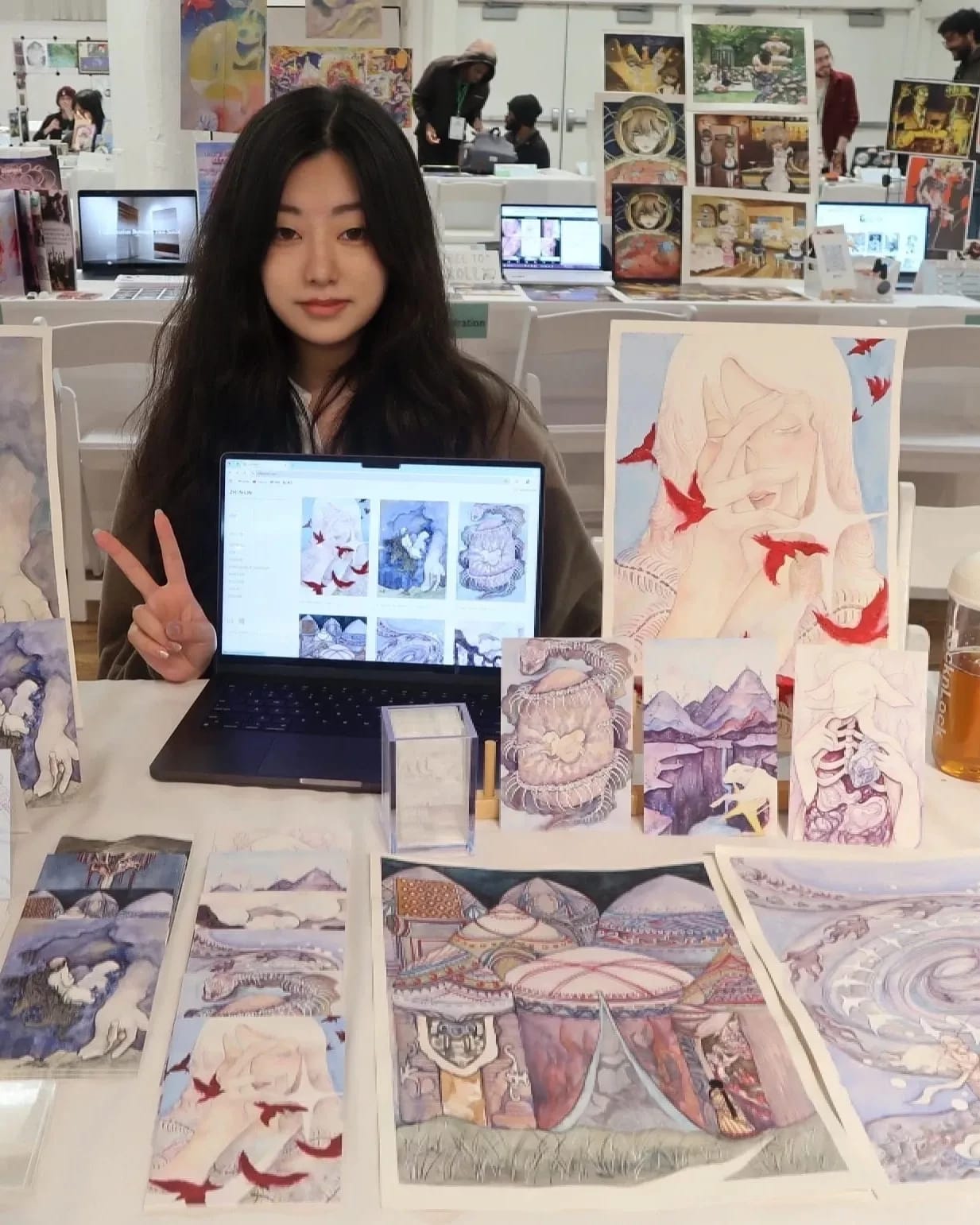 A girl poses at a table displaying a variety of artwork.