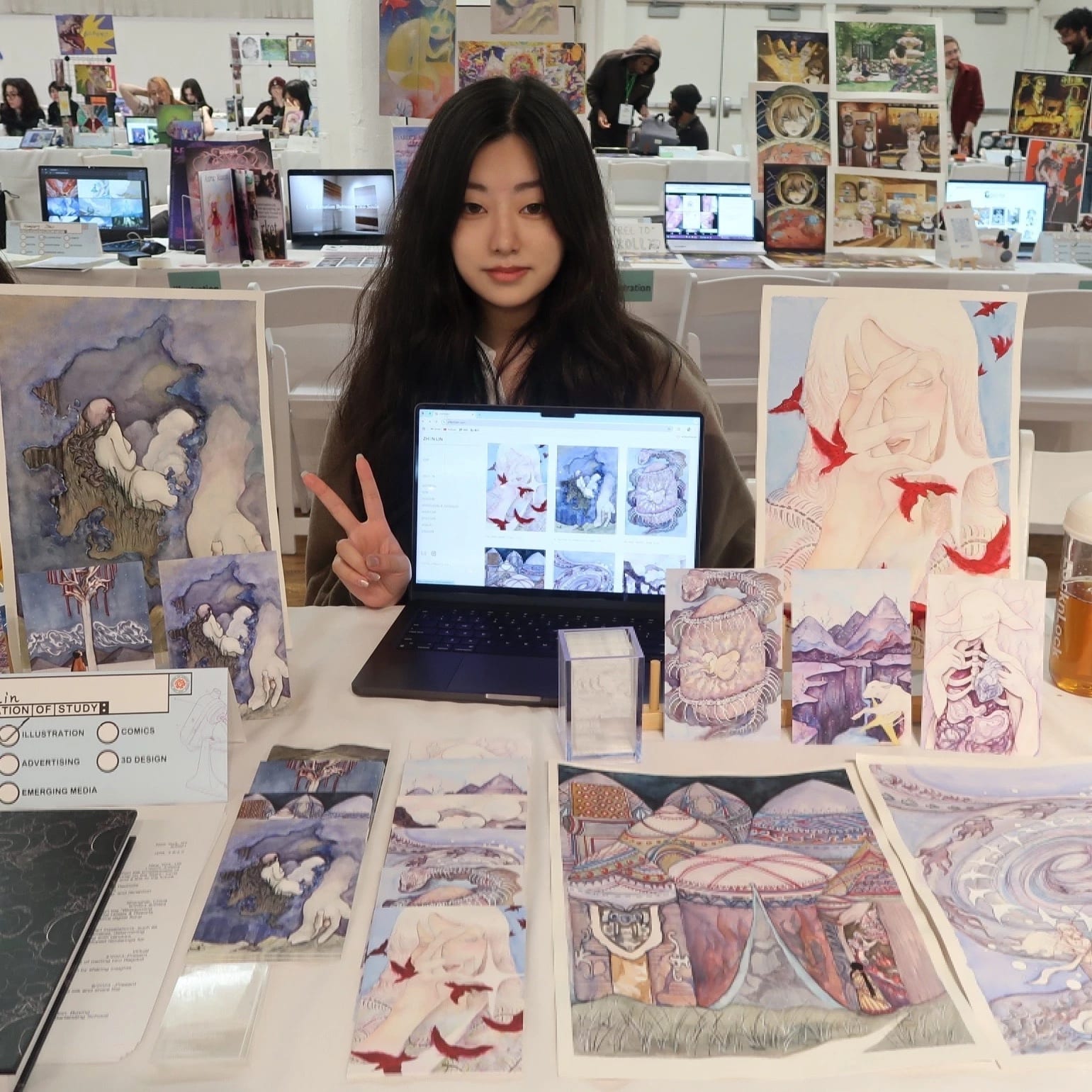A girl poses at a table displaying a variety of artwork.