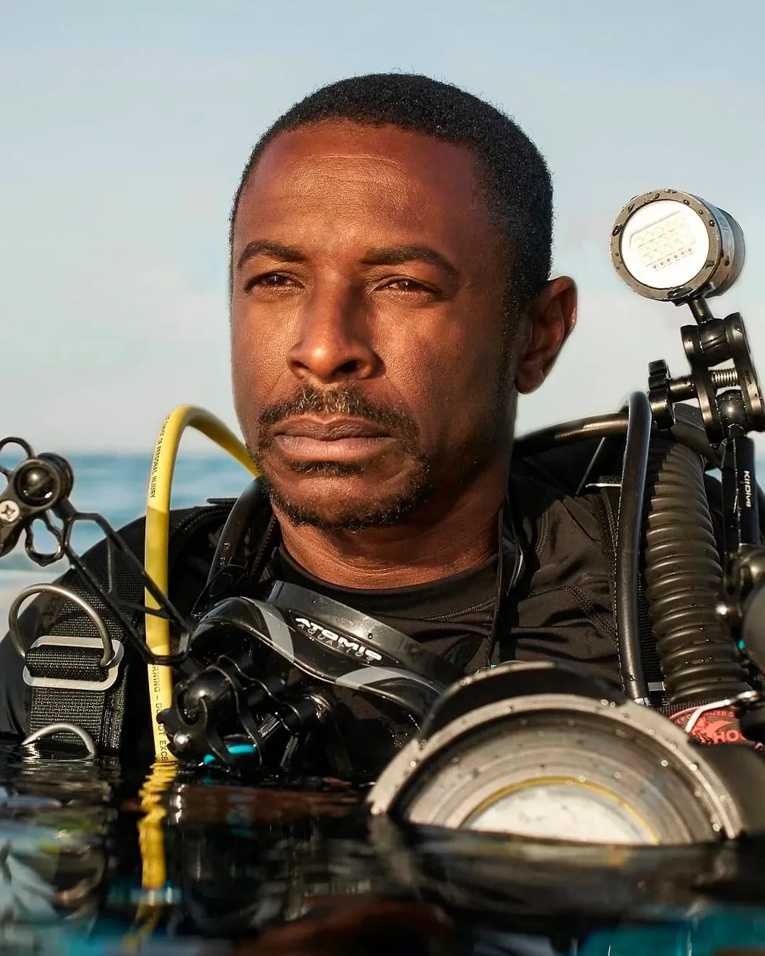 A portrait of a scuba diver in water. He has darker skin and short hair, is wearing a black top, and is equipped with numerous lighting and camera equipments strapped to his body. His body is submerged in water from the chest down.