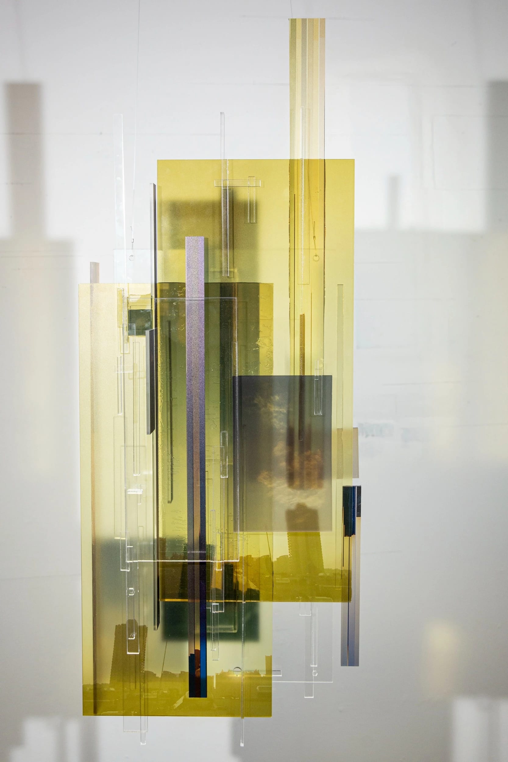 Black, yellow, clear and white acrylic plastic rectangles and squares with photos of a city and clouds arranged into a wall sculpture, casting shadows on the wall.