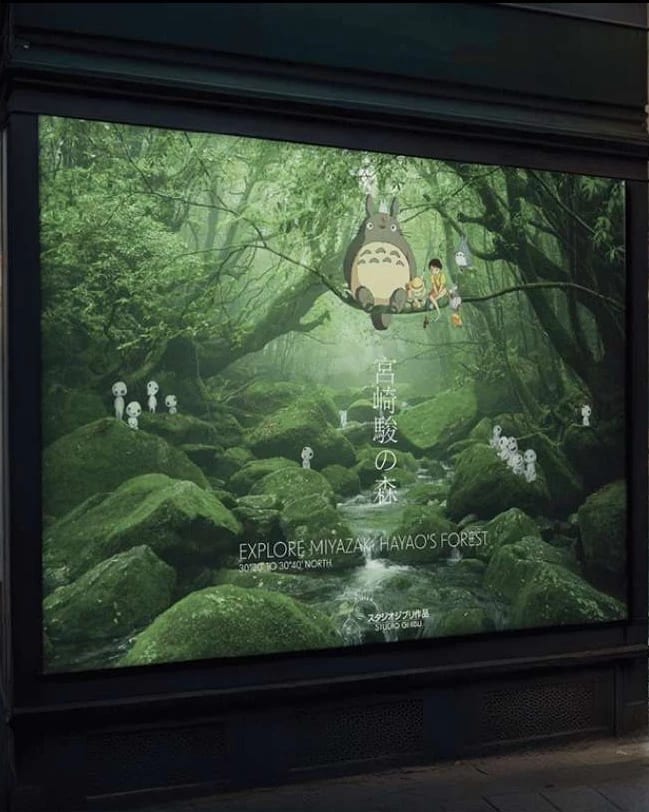 A mock-up of an advertising billboard on a newsstand; a verdant forest with whimsical creatures sitting on a tree branch and says "EXPLORE MIYAZAKI HAYAO'S FOREST."