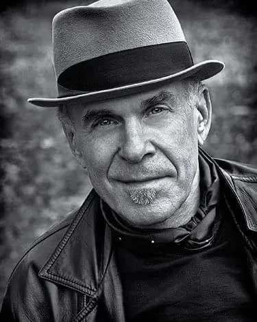A black and white photo portrait of the photographer Douglas Dubler. He has light skin, is wearing a fedora & a leather jacket over a black top with a silk scarf around his neck.