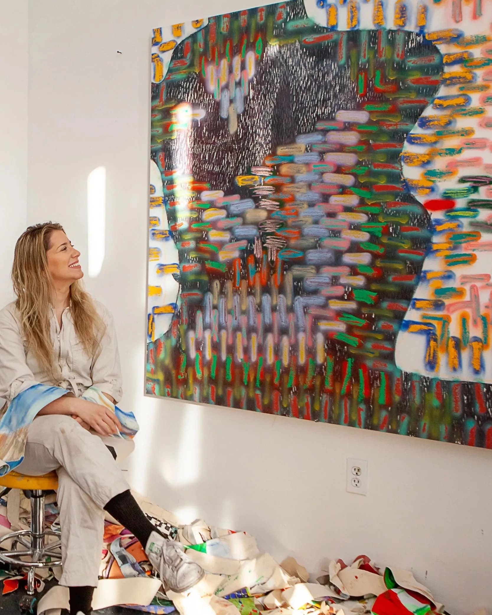 A blonde white woman wearing a taupe work jumpsuit smiles looking at an abstract painting while sitting in an artists' studio.