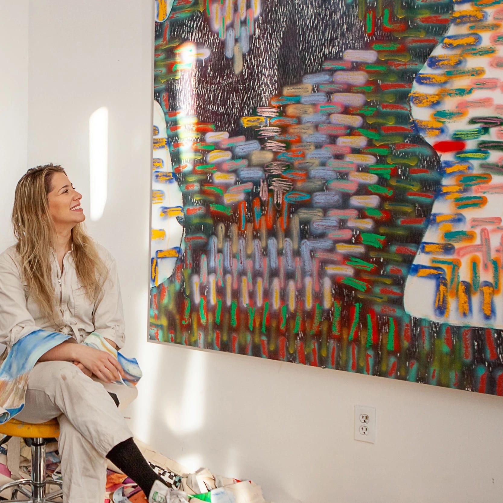 A blonde white woman wearing a taupe work jumpsuit smiles looking at an abstract painting while sitting in an artists' studio.