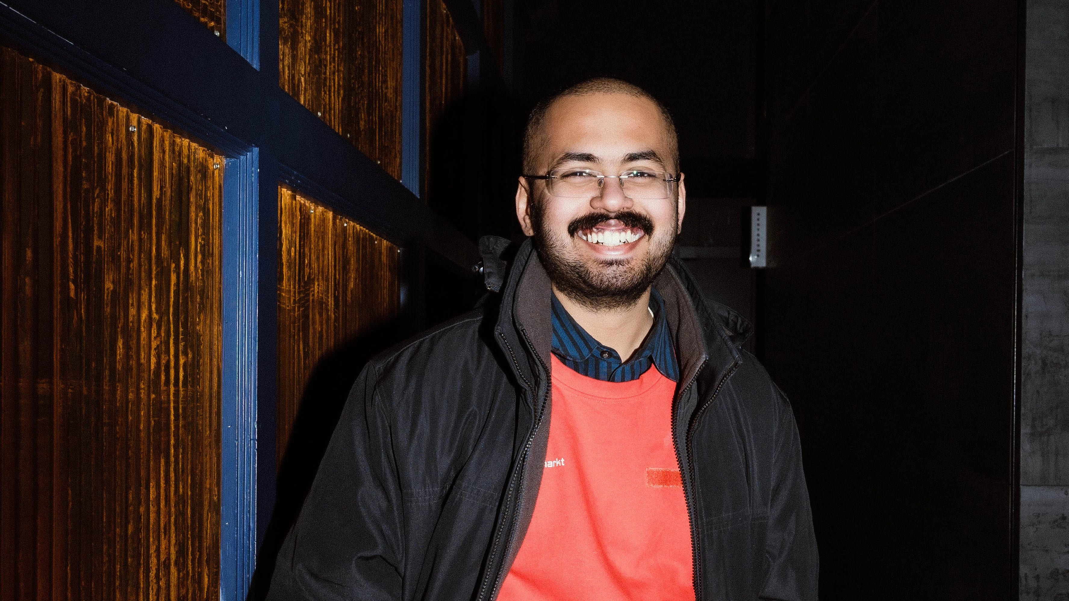 A man with a buzz cut with a dark mustache smiles at the camera wearing a red shirt and dark jacket.