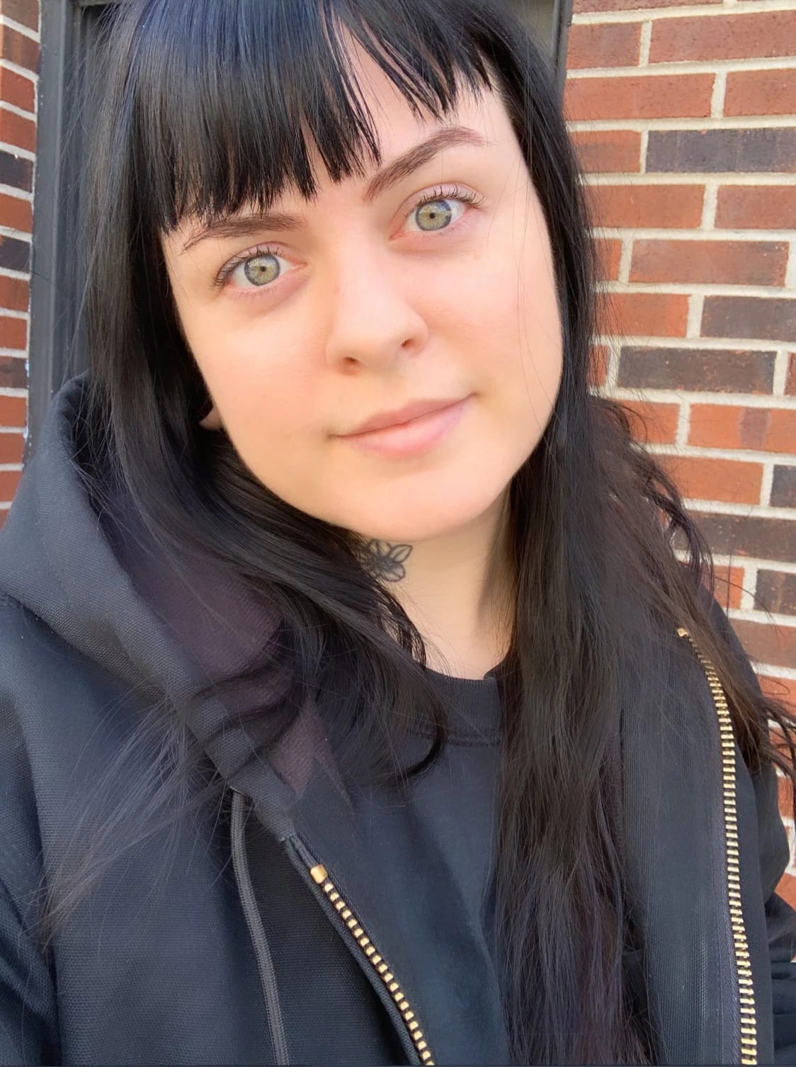 A headshot of Erin M. Riley. Erin is wearing a black shirt and black jacket. She is looking into the camera and smiling. In the background is a brick wall.