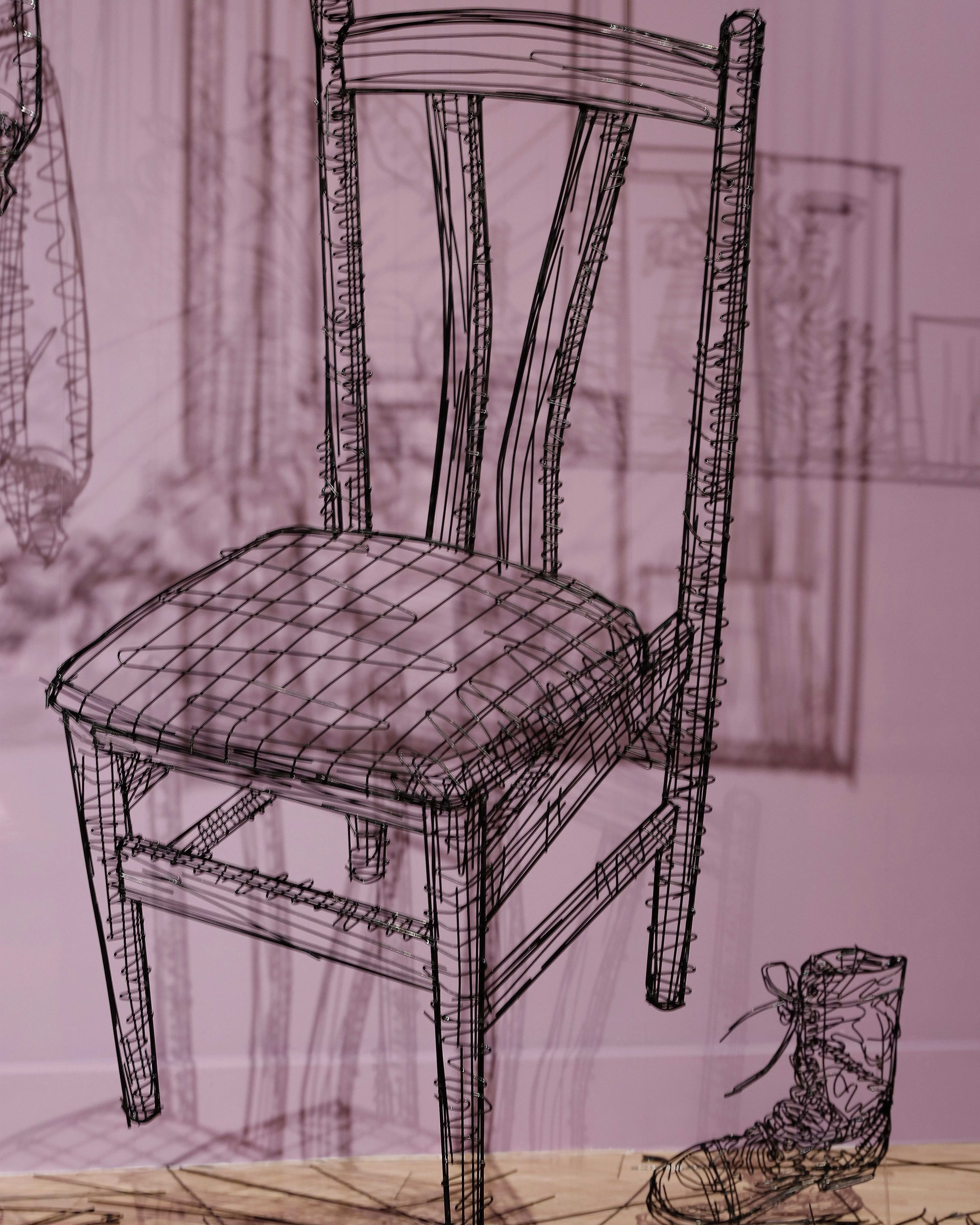 Close up of a sculpture made out of black wire depicting a wooden chair with a small boot next to it.