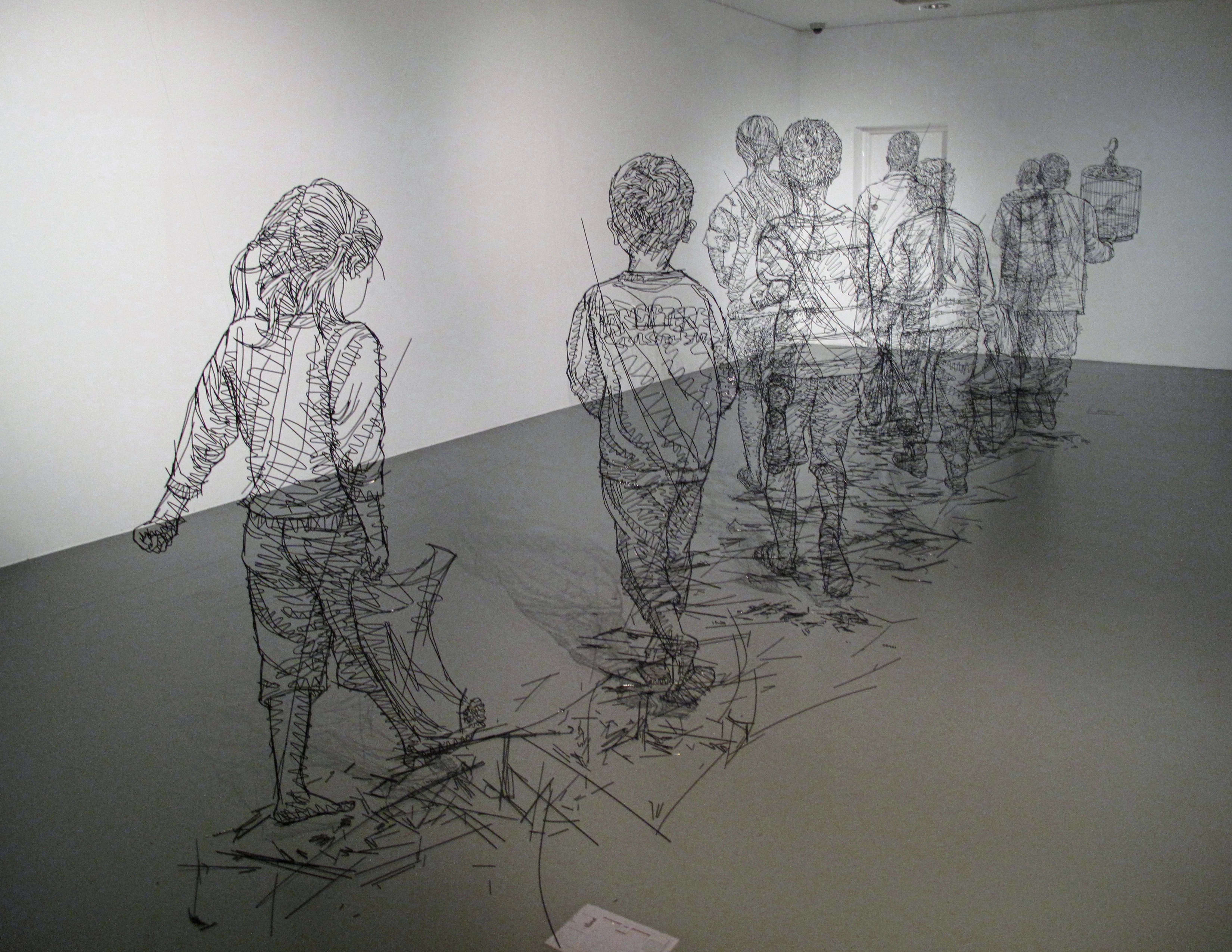 Sculptures made in black wire of several large scale children walking in a line in a white room. 