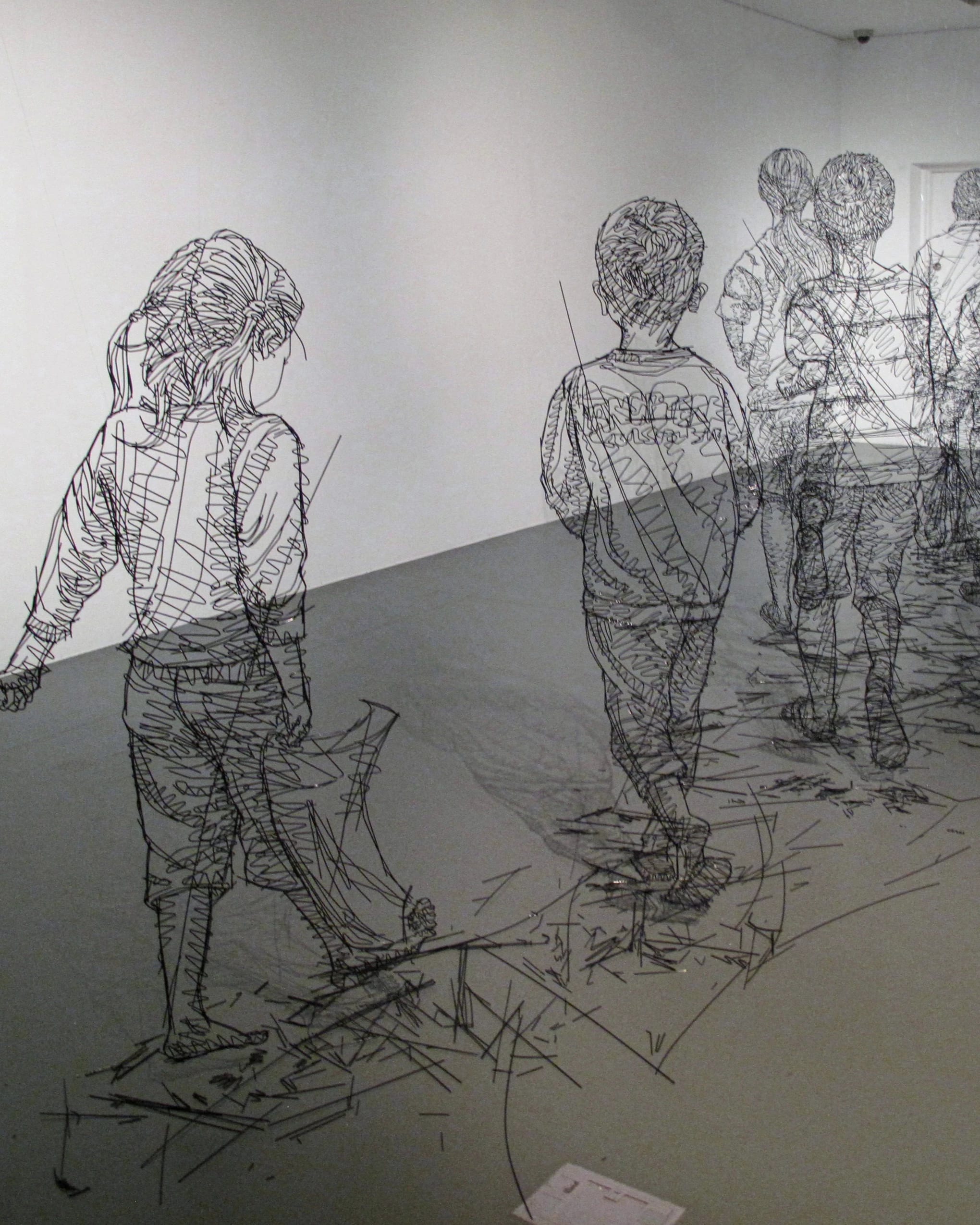 Sculptures made in black wire of several large scale children walking in a line in a white room. 
