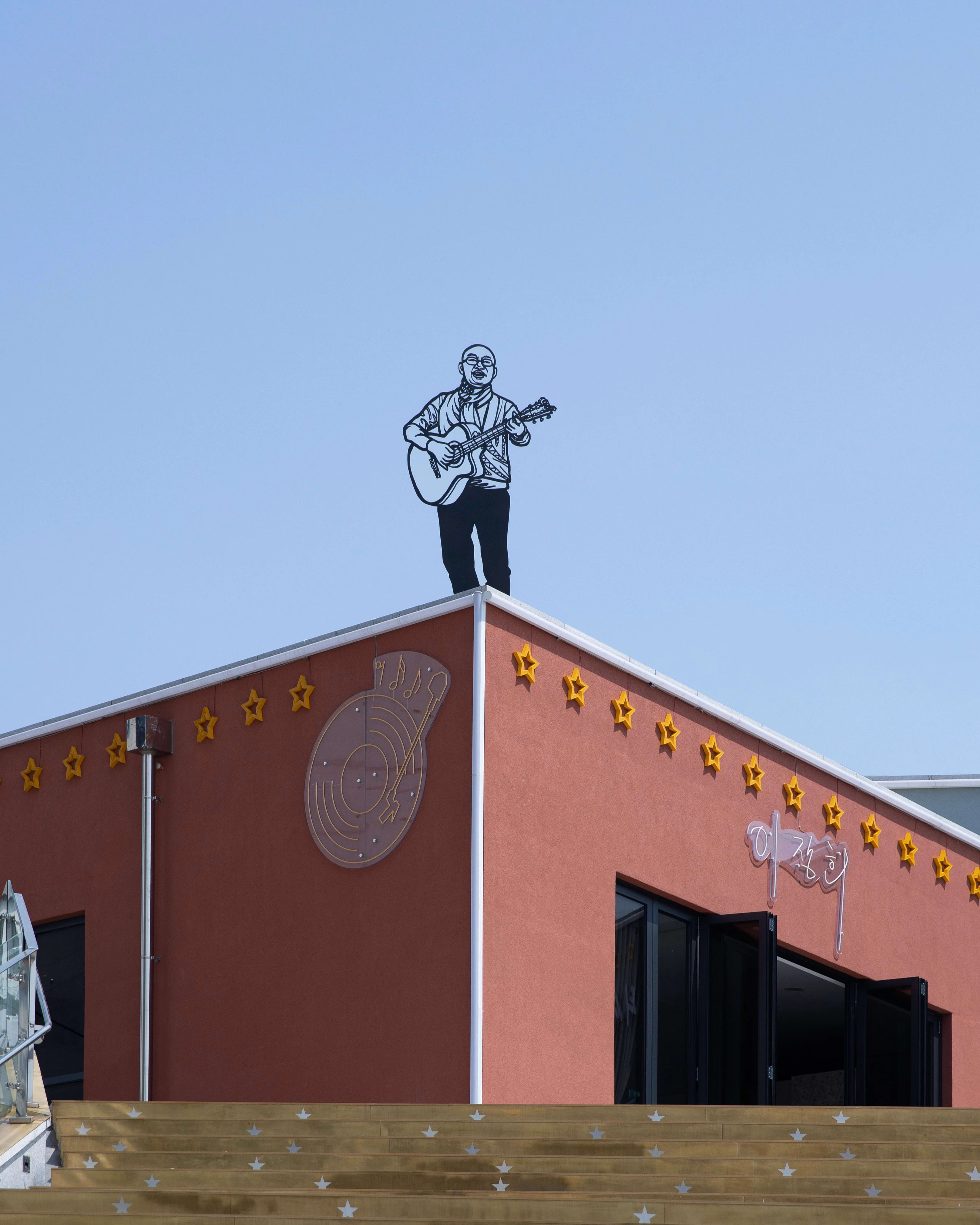 Sculpture made out of steel rods of a man playing guitar. Placed outside, on top of a short building.