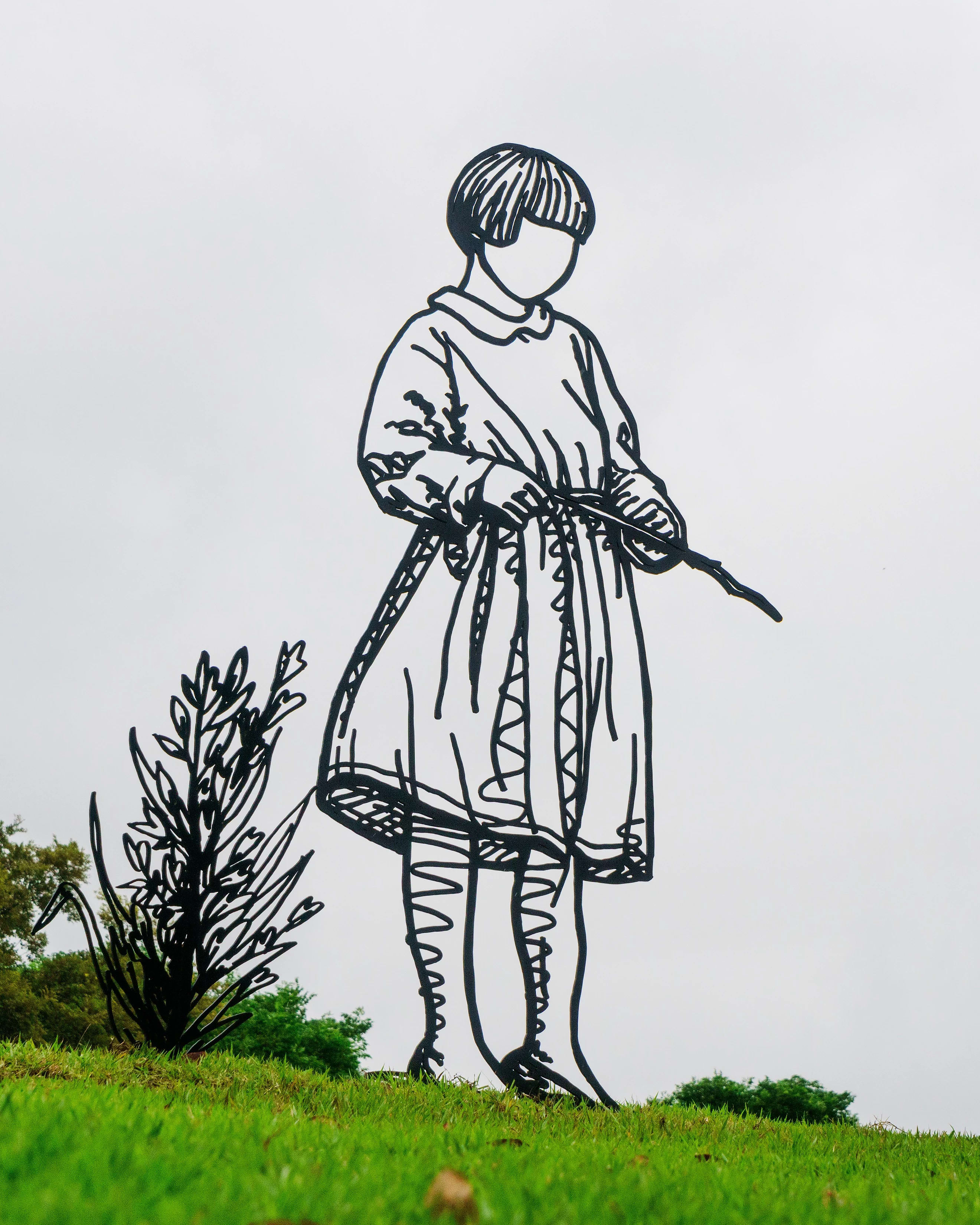 Sculpture made in steel rods on a little girl in a dress. The sculpture is set up outside, in a grassy place.