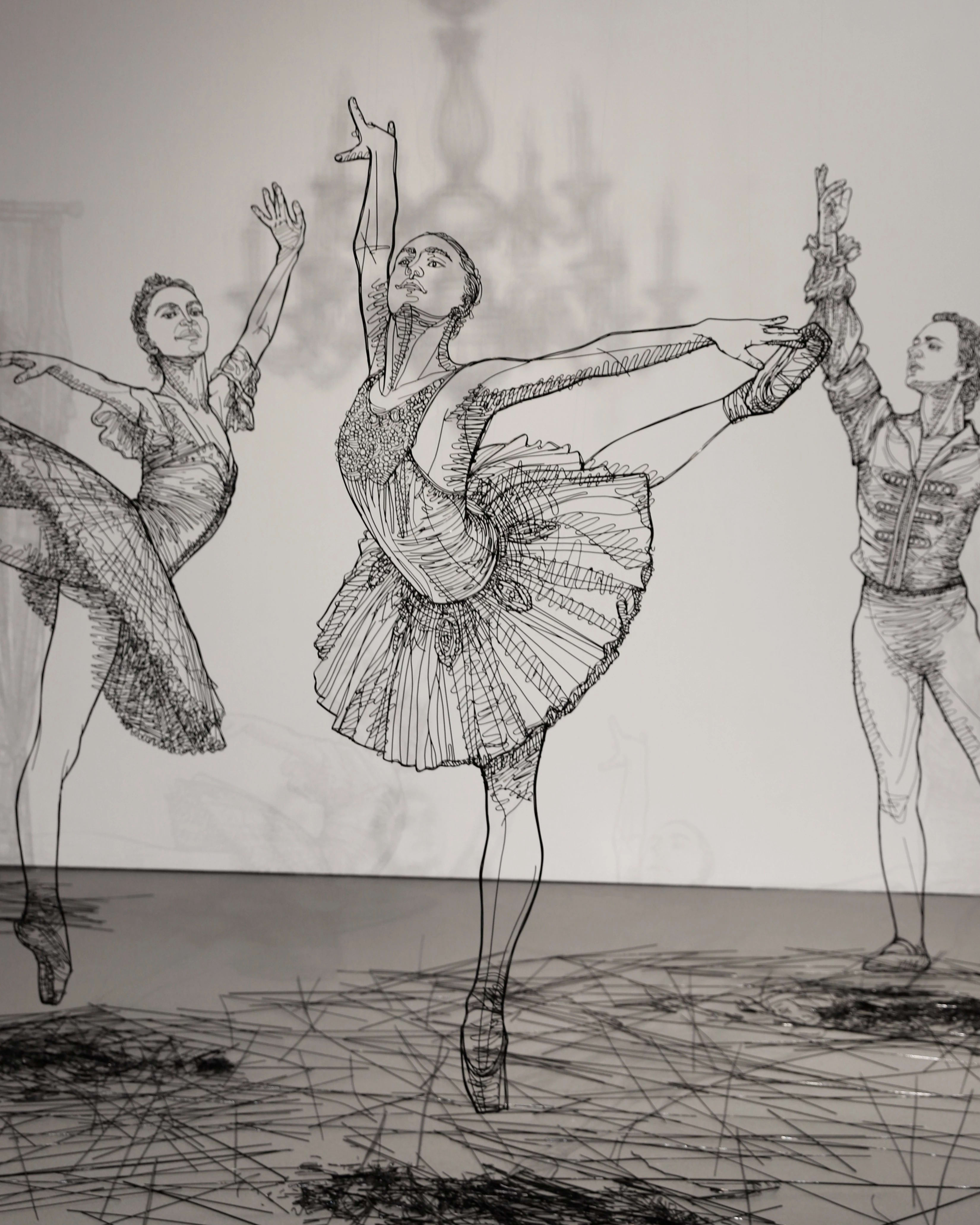 Close up of sculptures made in black wire of several large scale ballerinas dancing around a white room.