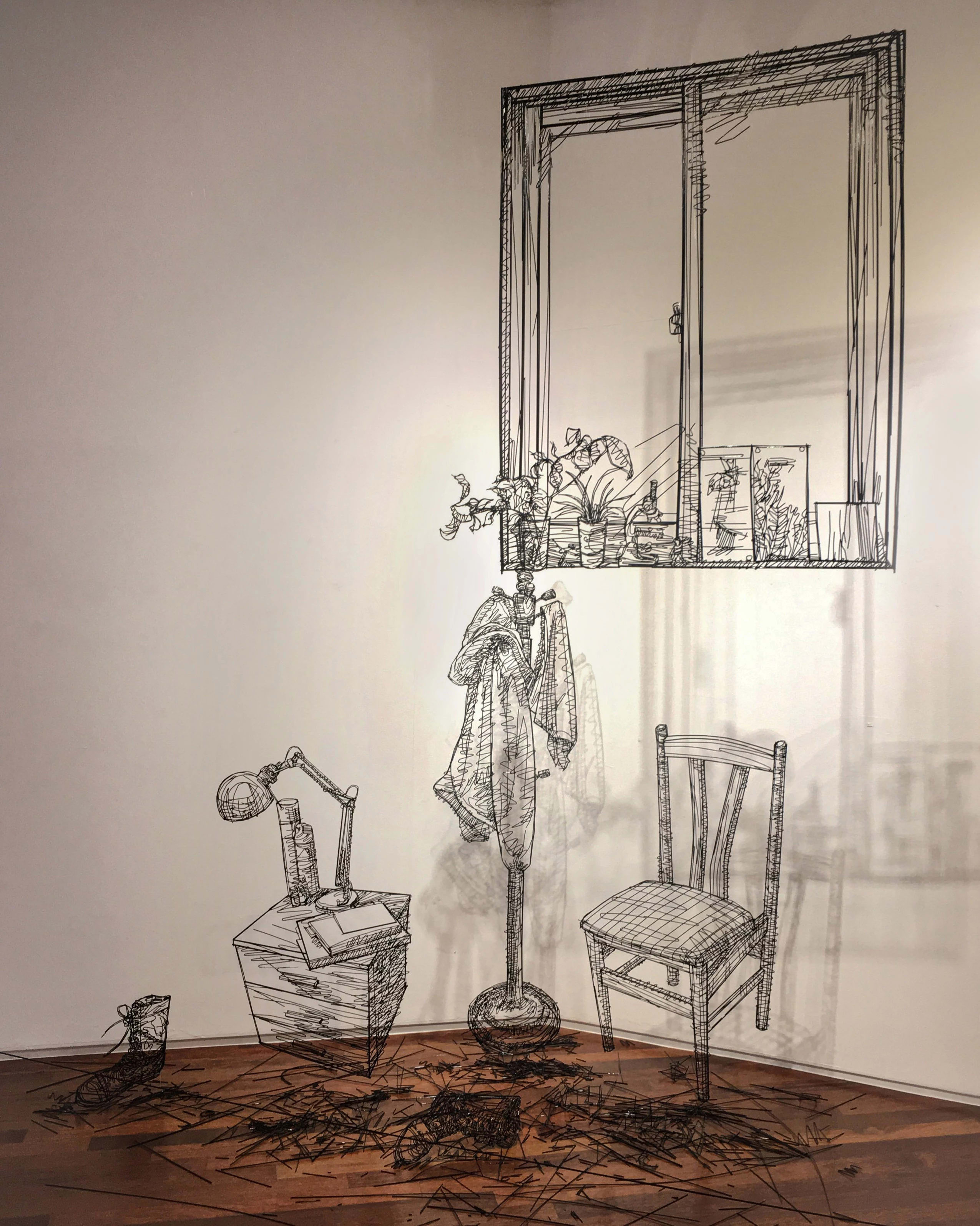 Sculptures made in black wire depicting the inside of a room. Including a wooden chair, a coat rack, a bedside table with a lamp and a window above them.