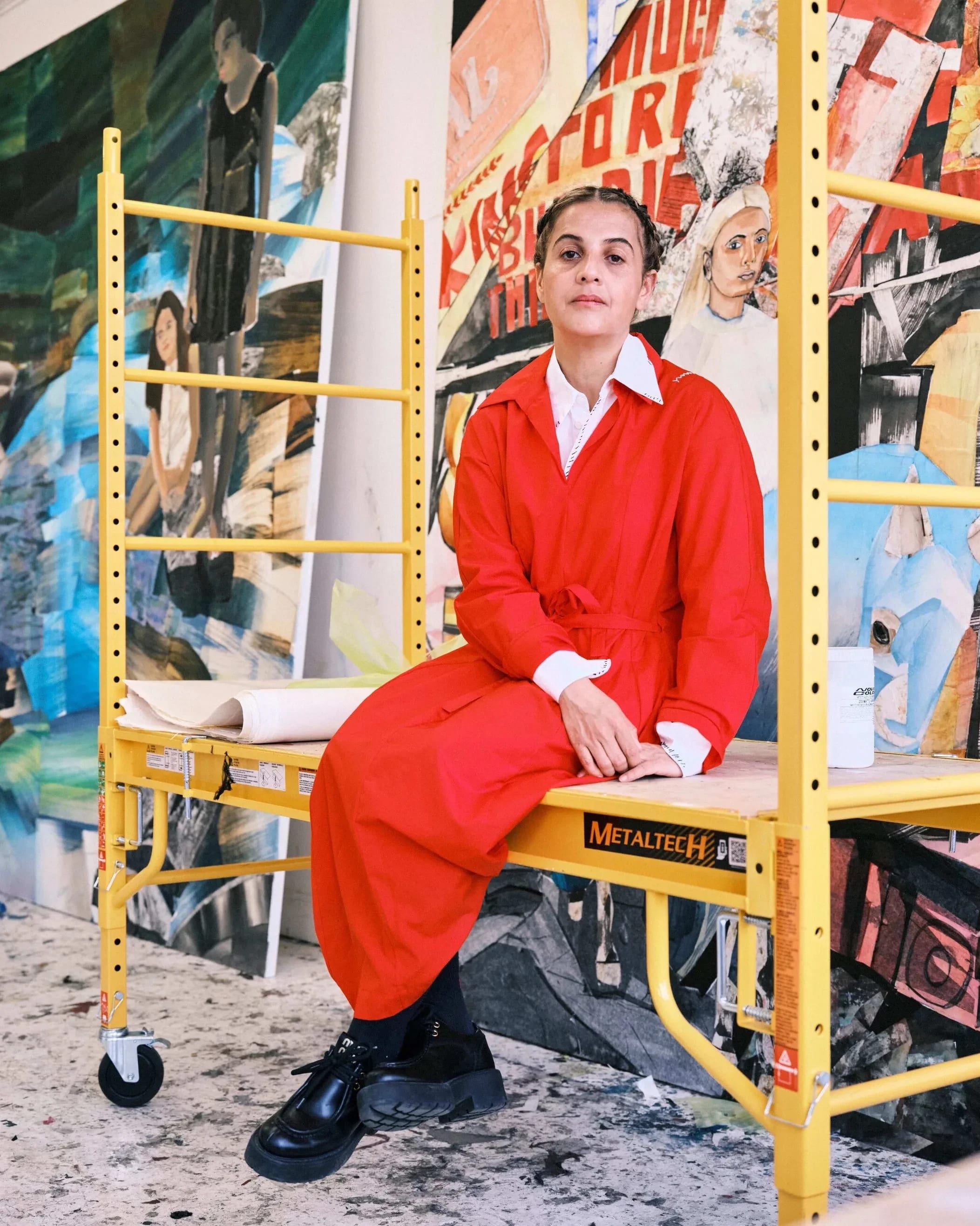 A woman in a bright red jumpsuit sits on a yellow metal frame on wheels in a studio.