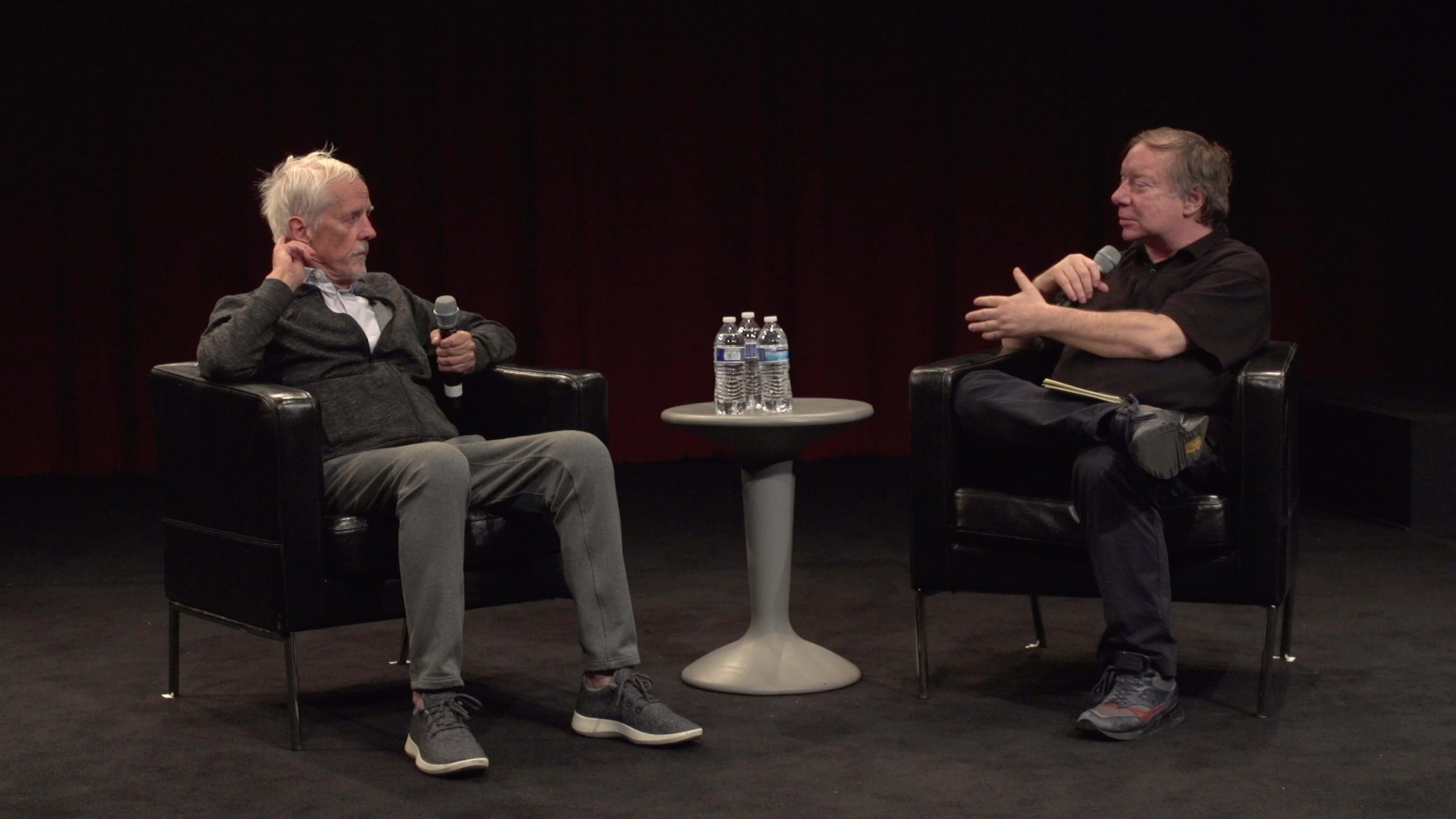 A color photograph of two people sitting in chairs on a carpeted floor, a table holding three unopened bottles of water between them. Each person is holding a microphone and the person on the right is gesturing and talking while the person on the left listens.
